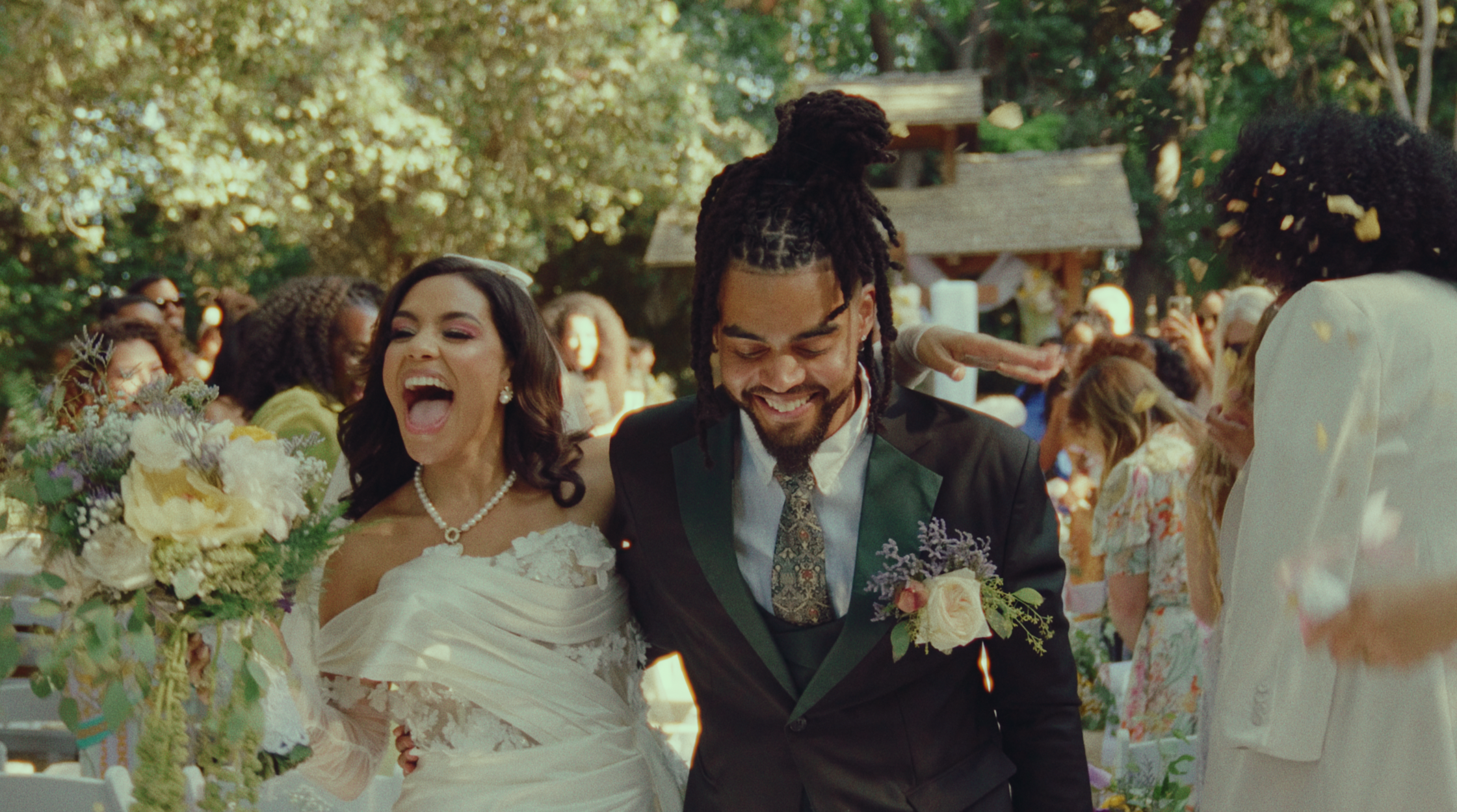 A bride in a white dress and a groom in a black suit walk happily together at their outdoor wedding, surrounded by guests and floral arrangements.
