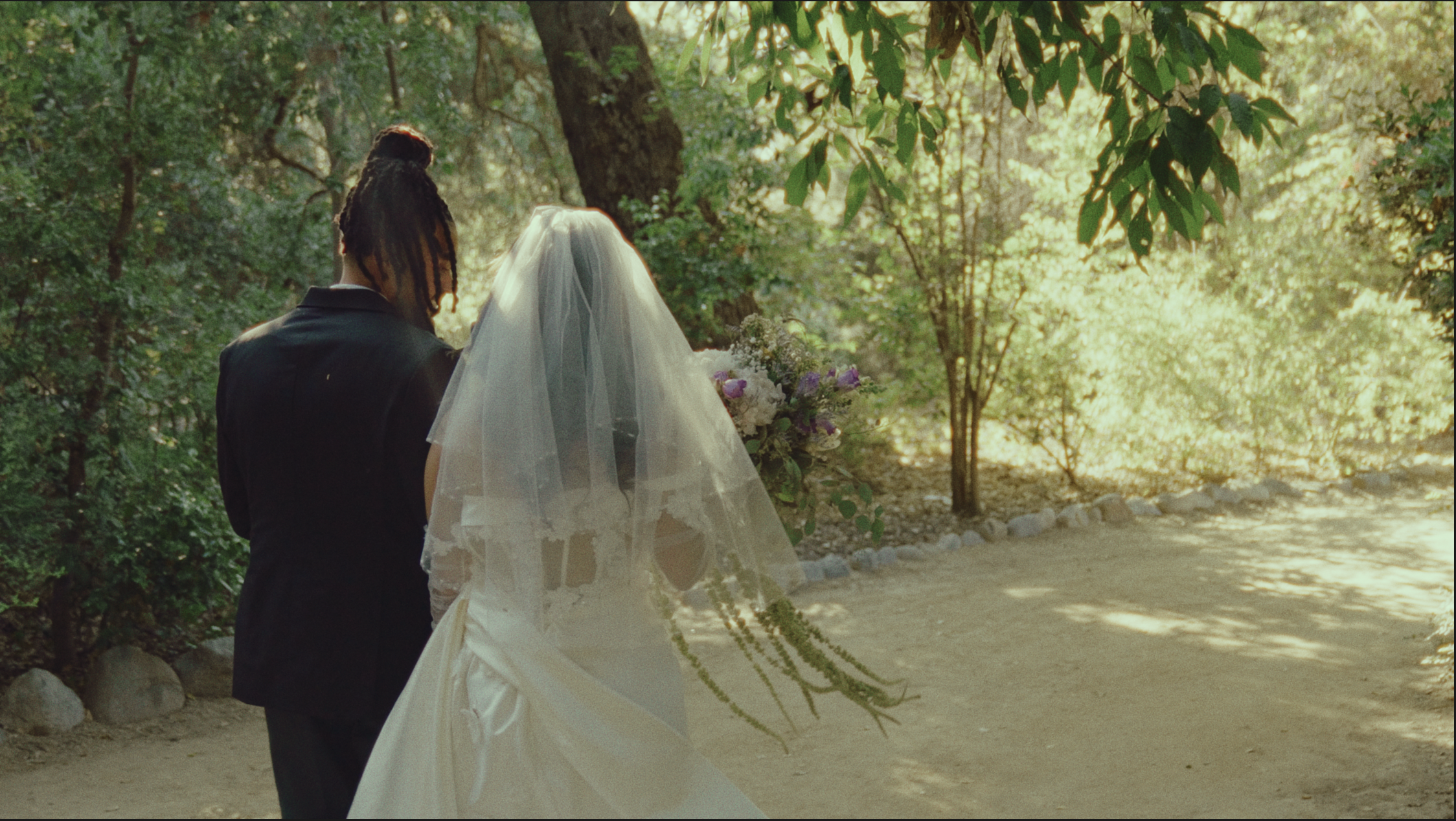 A bride and groom stand together outdoors surrounded by trees and sunlight. The bride wears a white wedding dress and veil, holding a bouquet of flowers. The groom wears a dark suit.