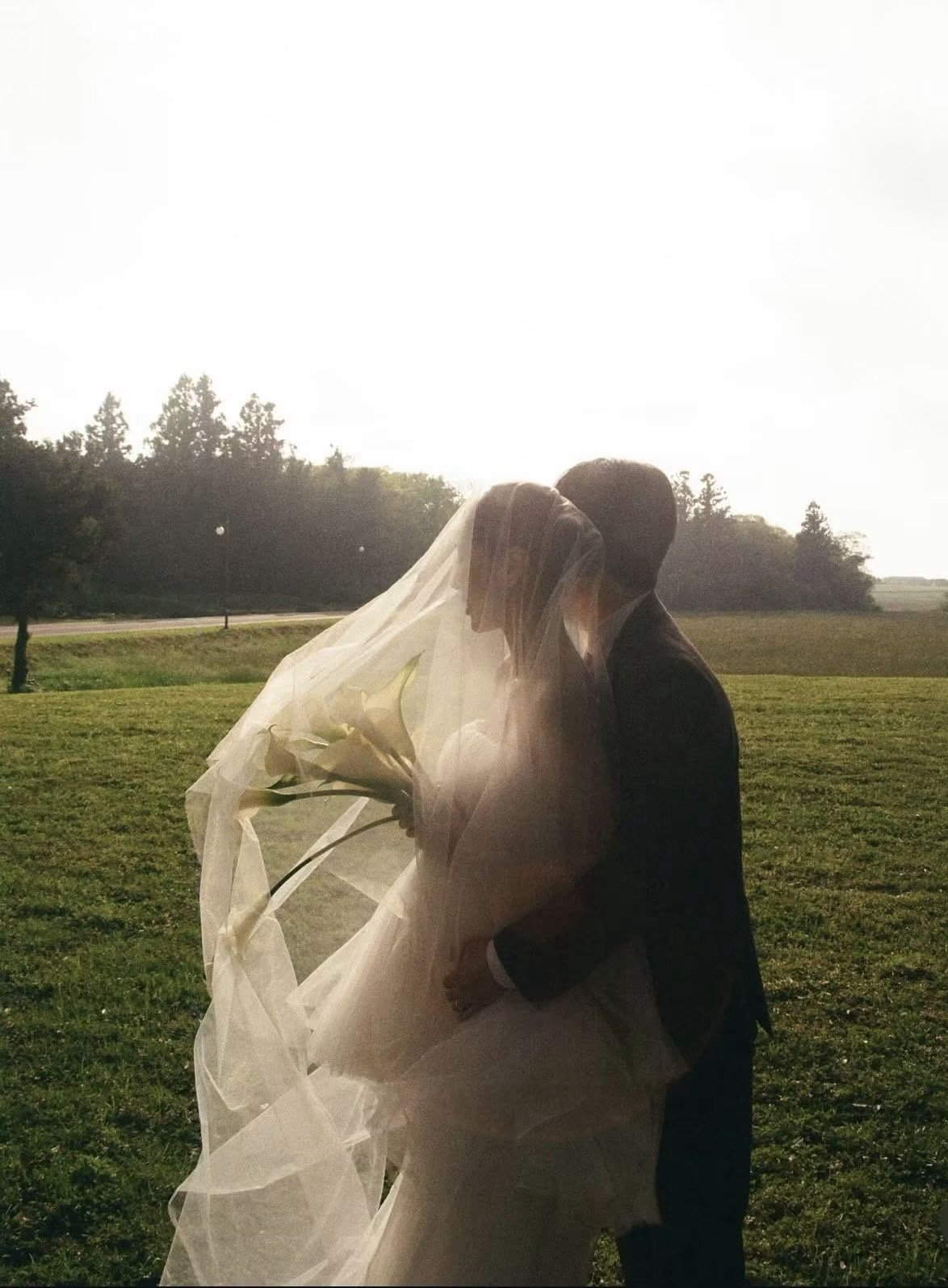 A couple dressed in wedding attire sharing an intimate moment outdoors during sunset, with the bride holding a bouquet and both veiled.