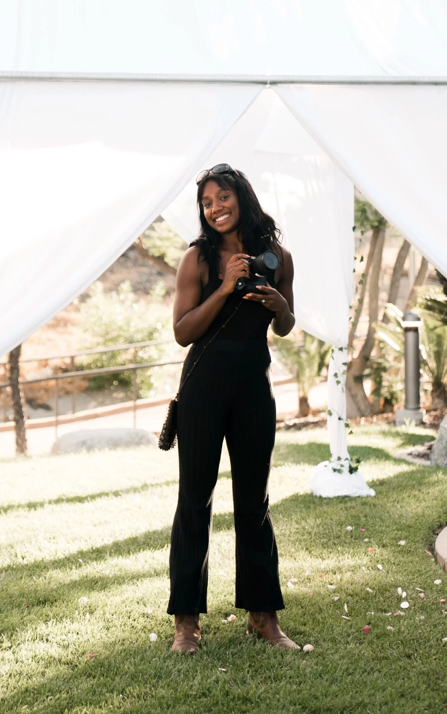 A woman standing on grass holding a camera, smiling, with sunglasses on her head and a black outfit, in front of a white tent outdoors.