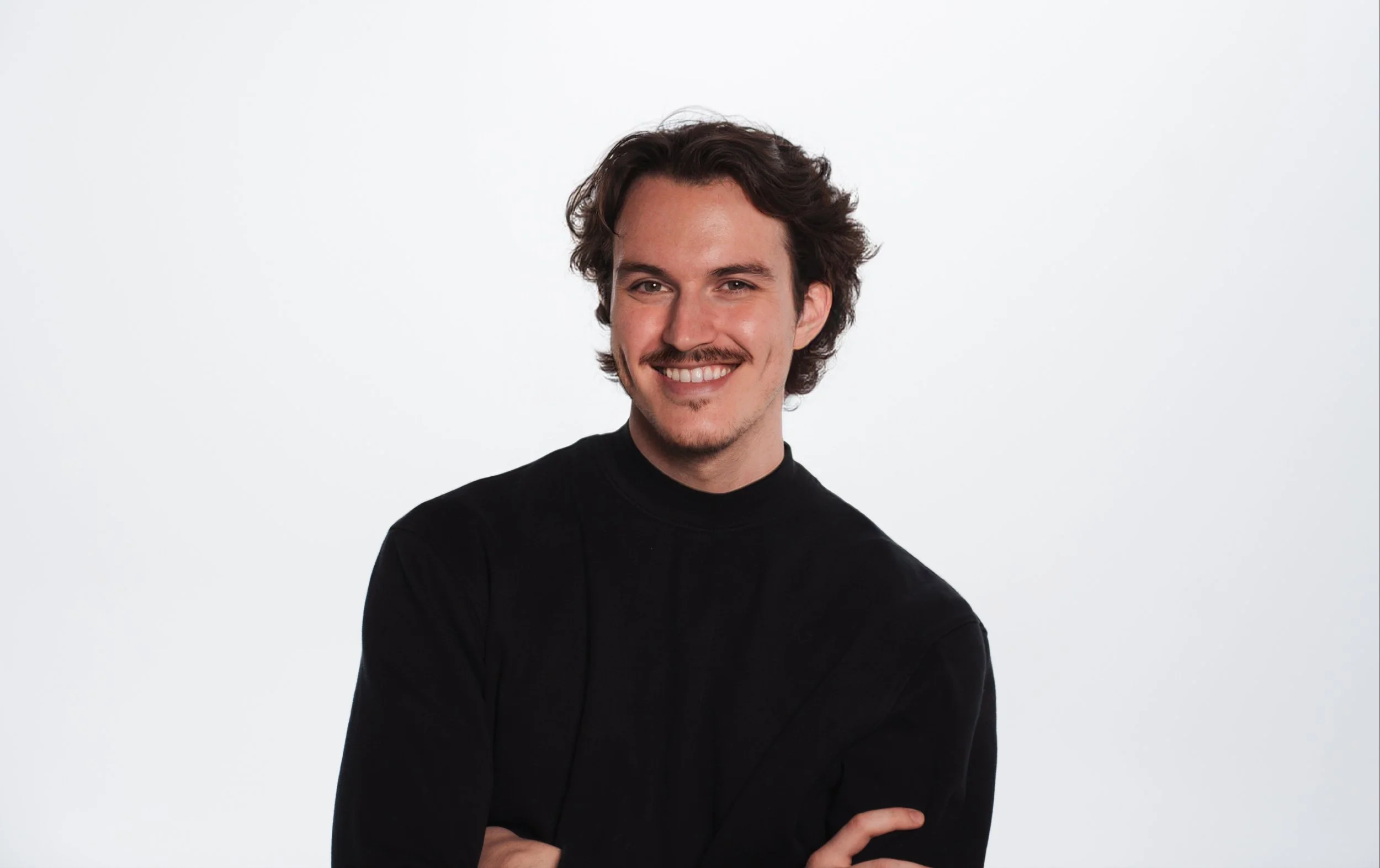A young man with curly hair, wearing a black shirt, smiling with arms crossed against a plain white background.