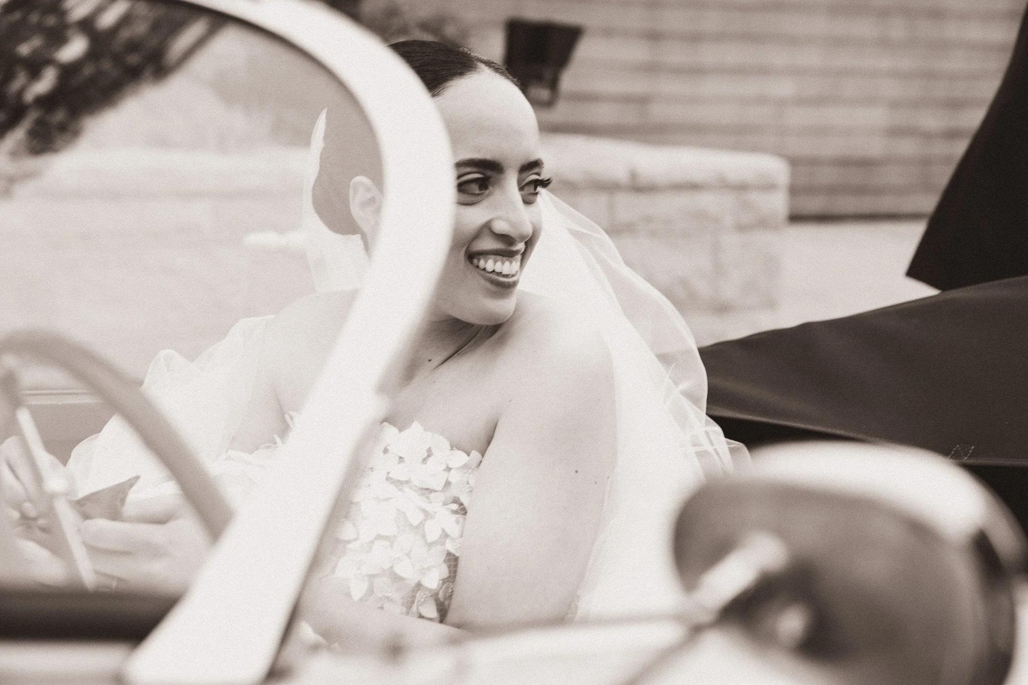 A woman in a wedding dress smiling inside a vintage car during a wedding ceremony.