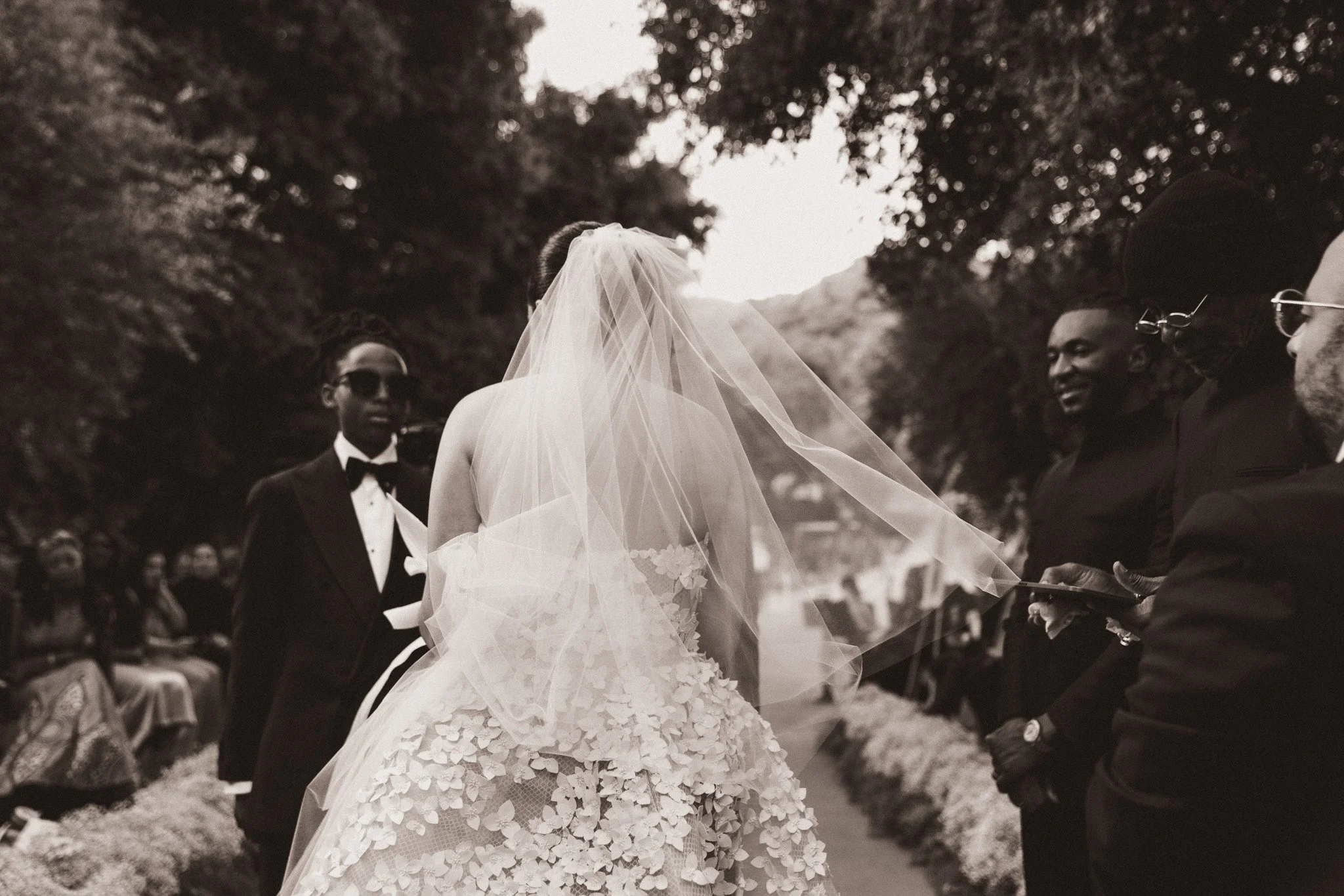 A woman in a wedding dress with a veil stands outdoors during a wedding ceremony, surrounded by friends and family. The women and men on her right and left are dressed in formal attire, with some wearing sunglasses. The setting appears to be a natura