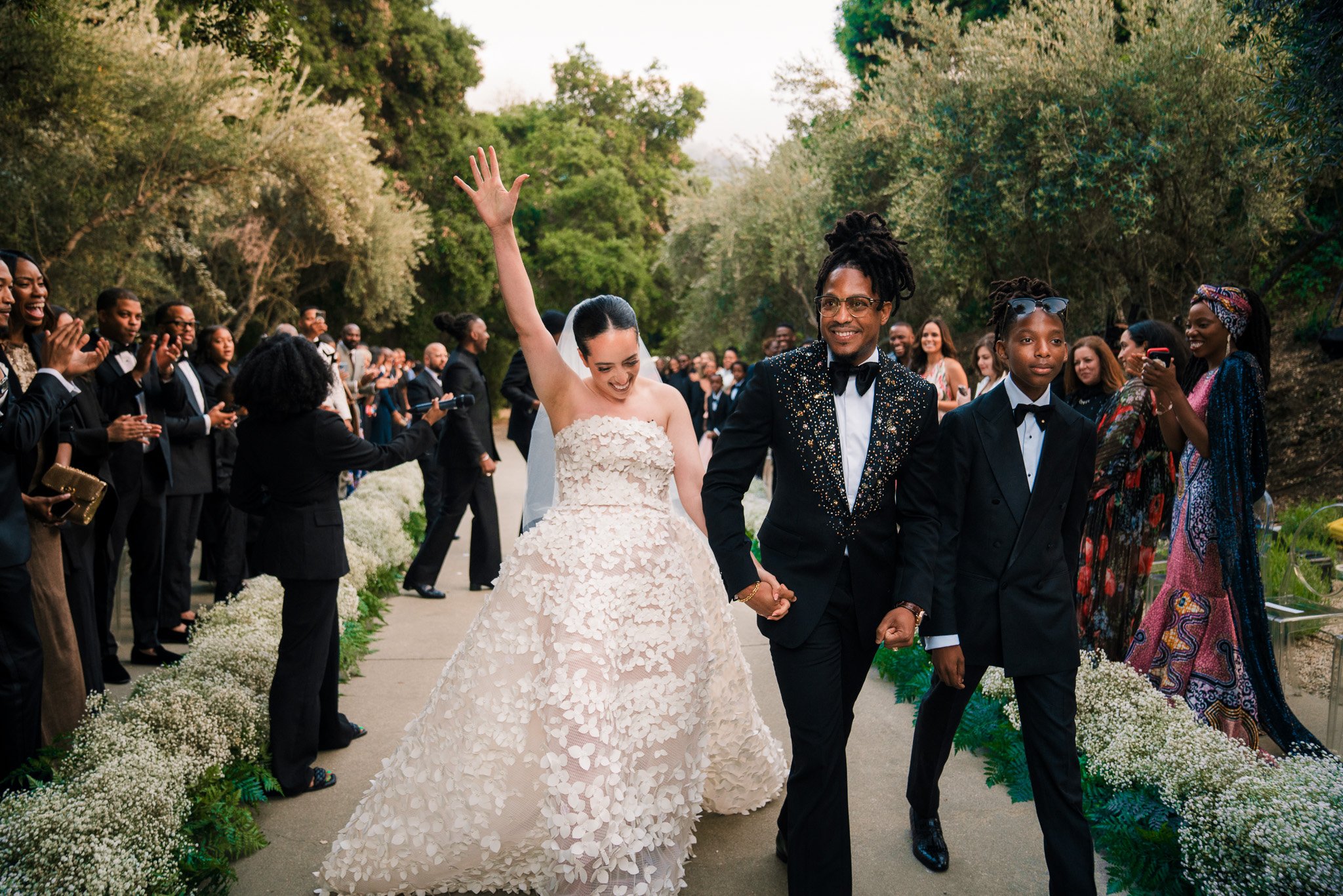 A newlywed couple walks down the aisle, holding hands. The bride is wearing a strapless wedding dress with floral details, waving happily. The groom is dressed in a black tuxedo with a bow tie, smiling. Guests on both sides are clapping and taking ph