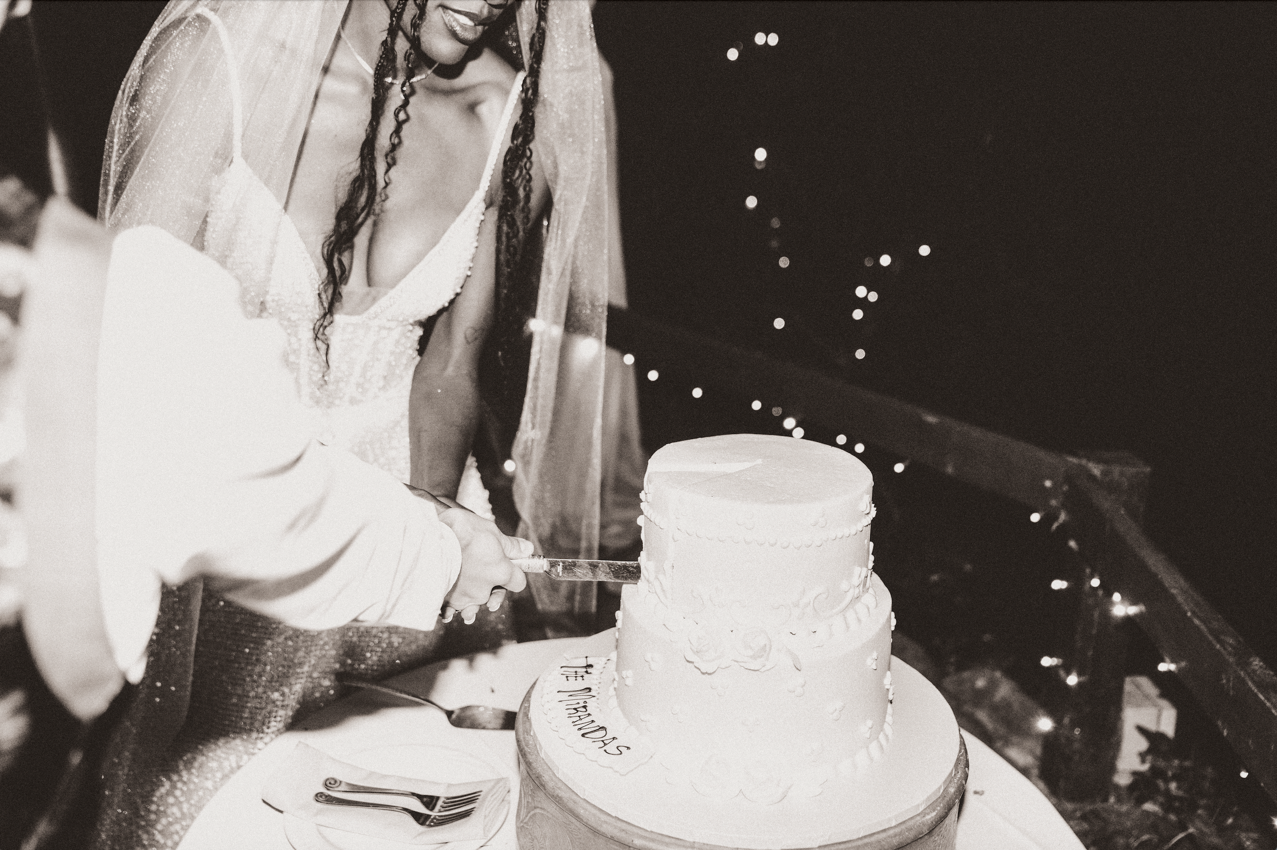 A bride and groom cutting a wedding cake at their reception. The bride is wearing a lace wedding dress and a veil, and the groom is wearing a light-colored suit. The wedding cake is three-tiered with decorative flowers and icing. The scene is decorat