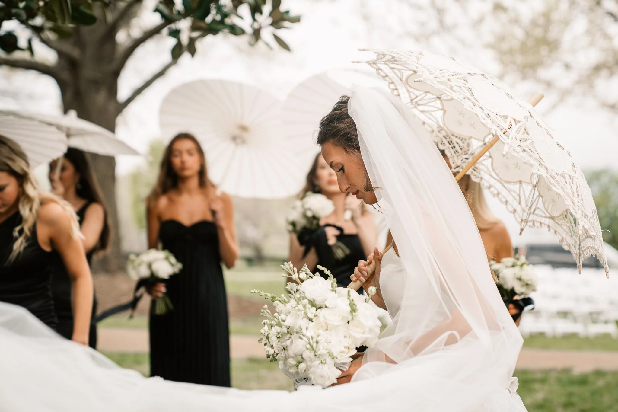 A bride in a wedding dress and veil holding a bouquet of white flowers, surrounded by bridesmaids holding similar bouquets and umbrellas, outdoors near a tree.
