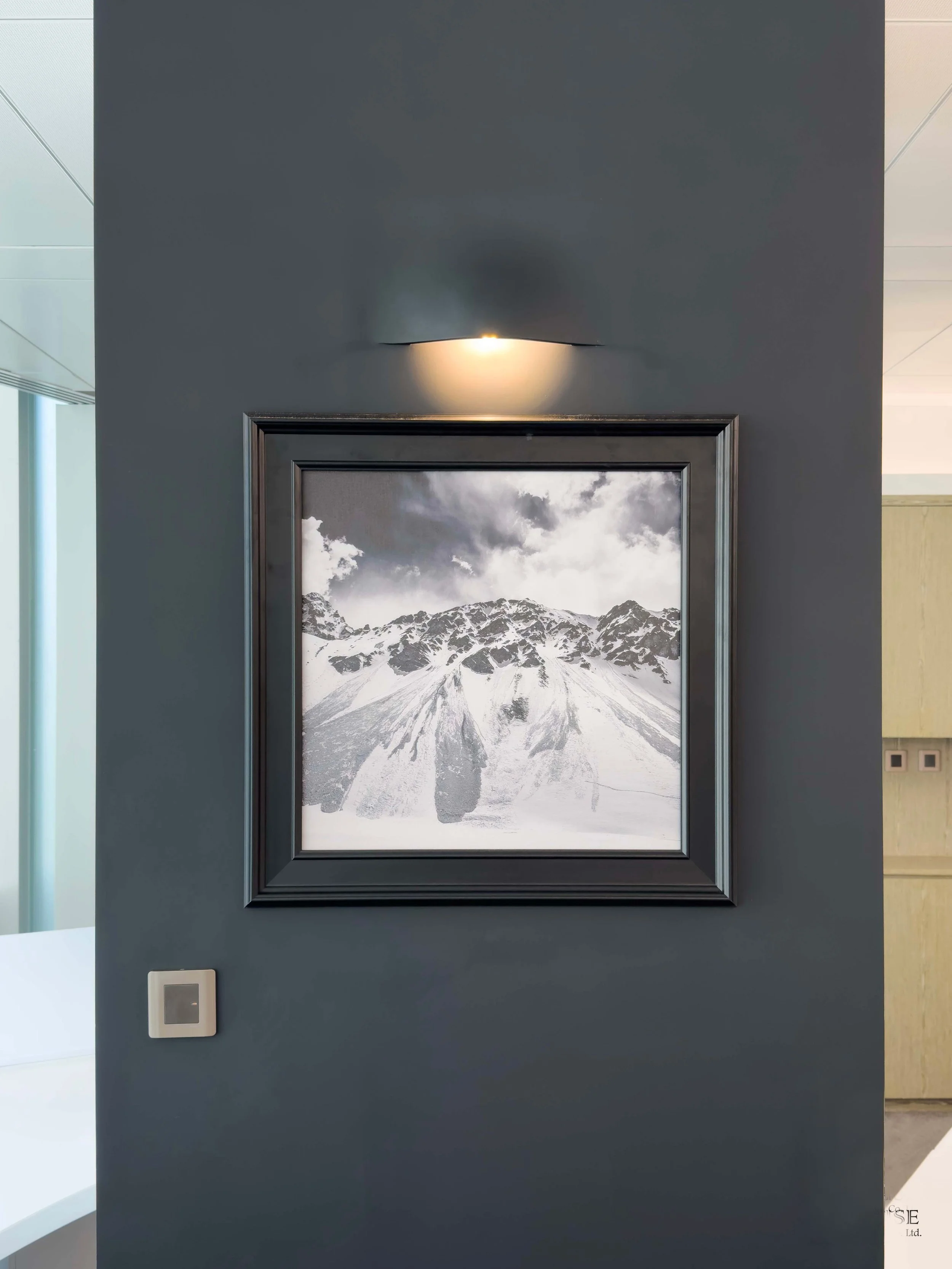 Black and white photograph of a snow-covered mountain with clouds in the sky, framed and hung on a dark gray wall, with a light above.