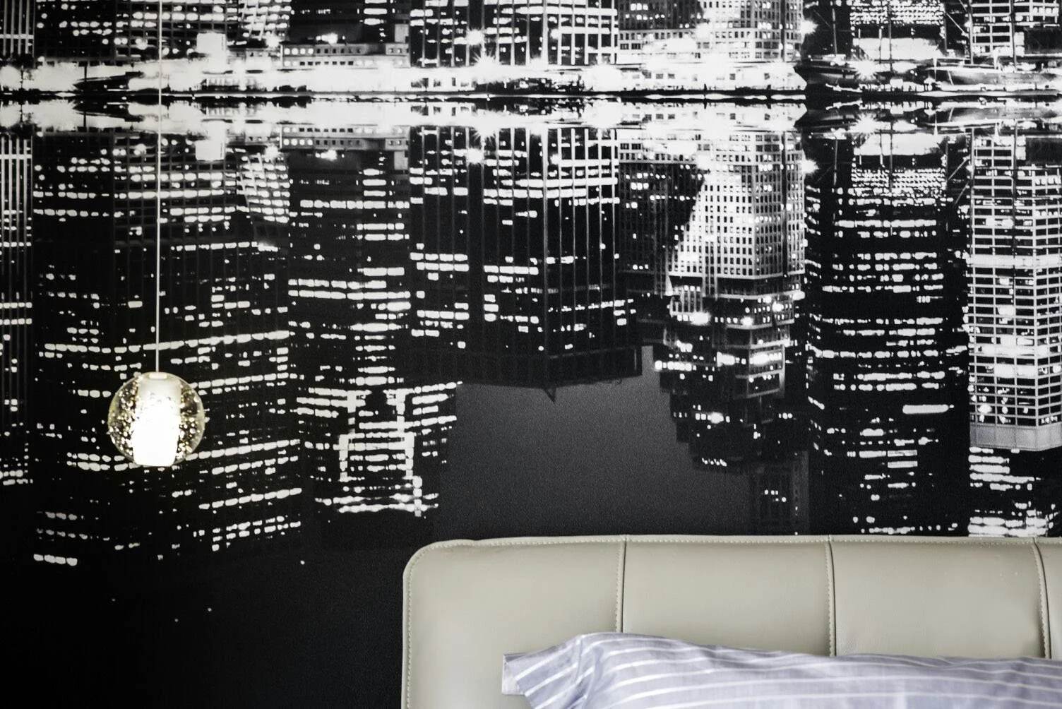 Bedroom with a large black and white cityscape mural of illuminated skyscrapers at night above a bed with a beige headboard and a striped pillow.