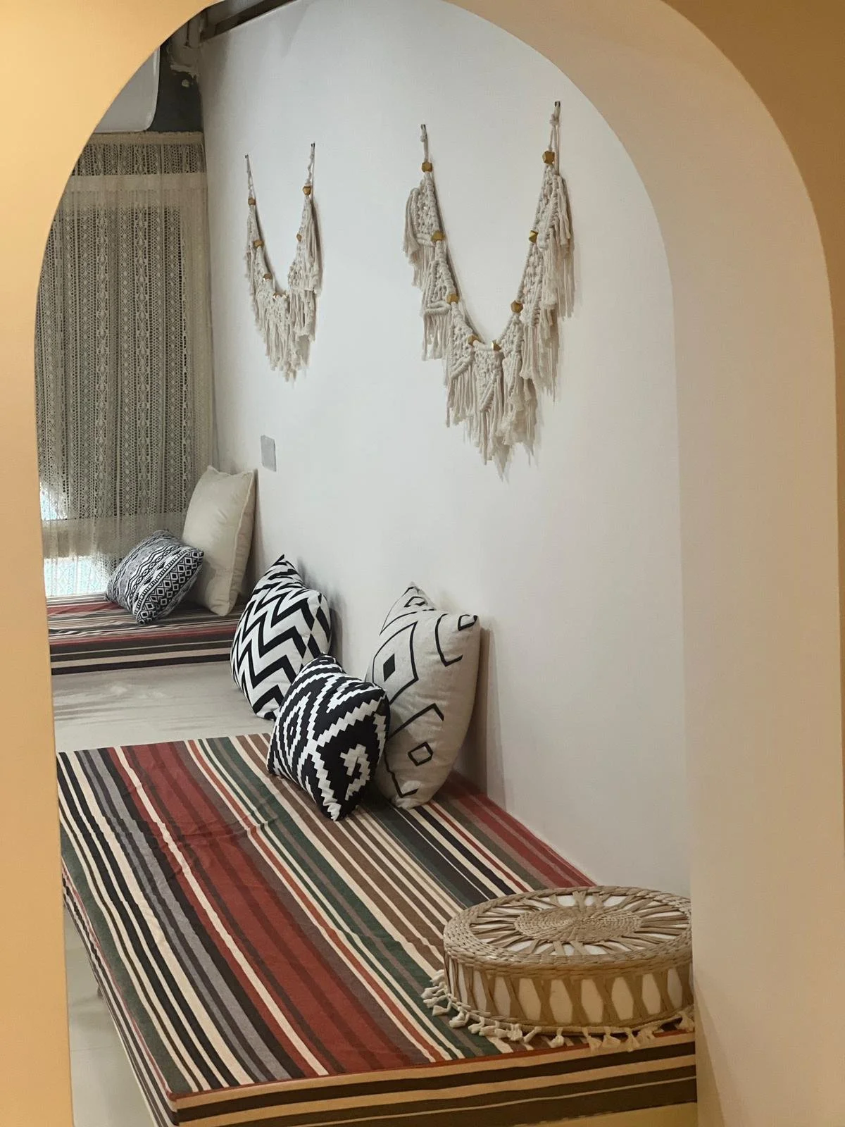 A cozy bed with striped bedding, black and white patterned pillows, and a woven rattan ottoman. The white wall behind the bed has three boho-style macrame wall hangings.