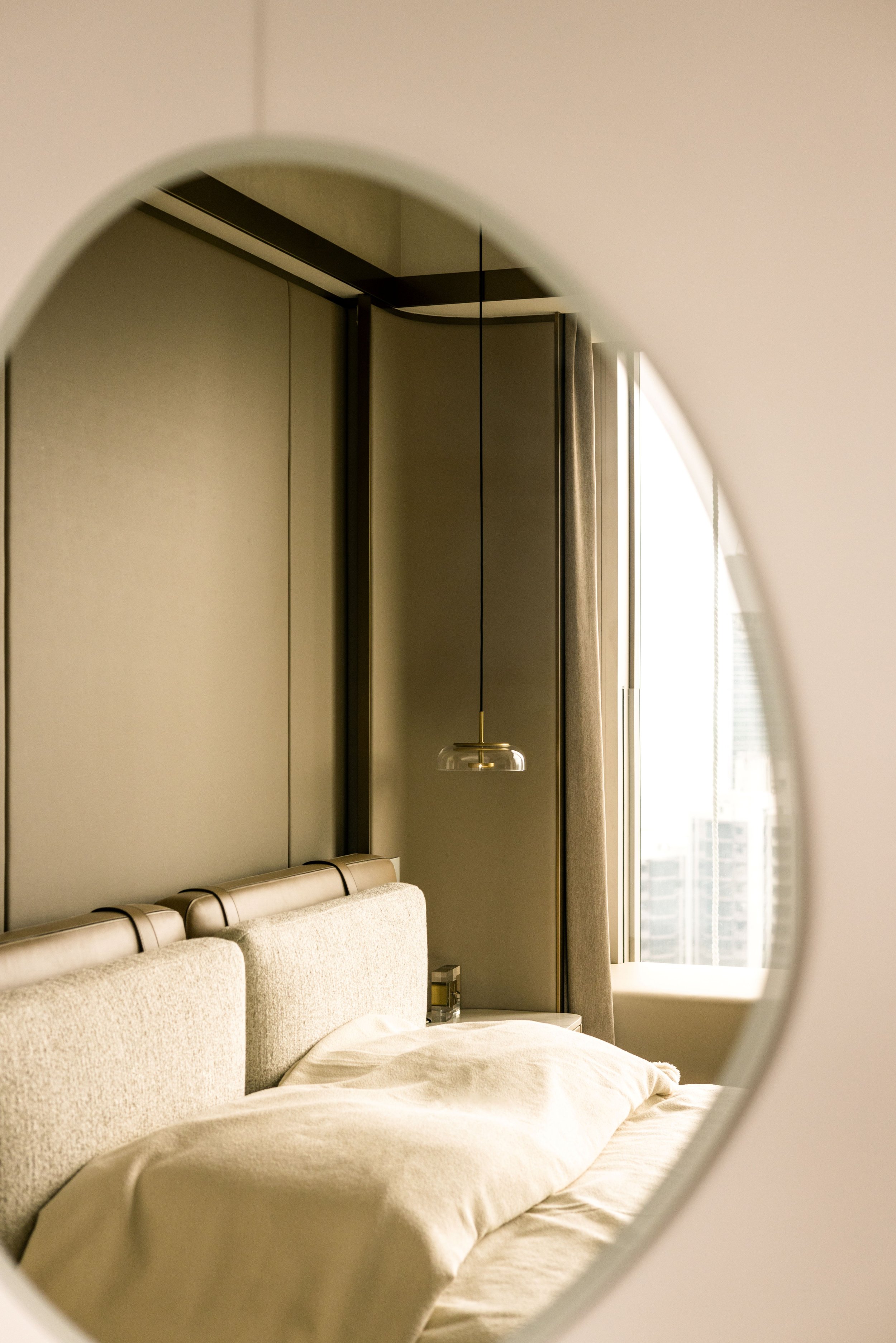 A hotel room with a beige upholstered bed, a bedside table with a glass, a modern pendant light, and large windows with curtains.