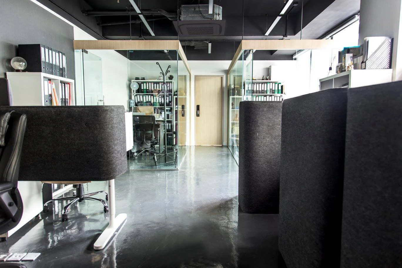 Modern office with glass-enclosed meeting rooms, white shelves with binders, black office chairs, and a polished concrete floor.