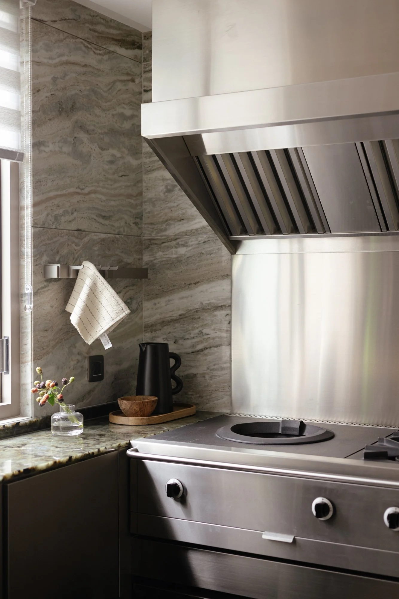 Modern kitchen with stainless steel stove and range hood, marble countertops, gray marble walls, window with blinds, black kettle, wooden bowl, and small vase with flowers.