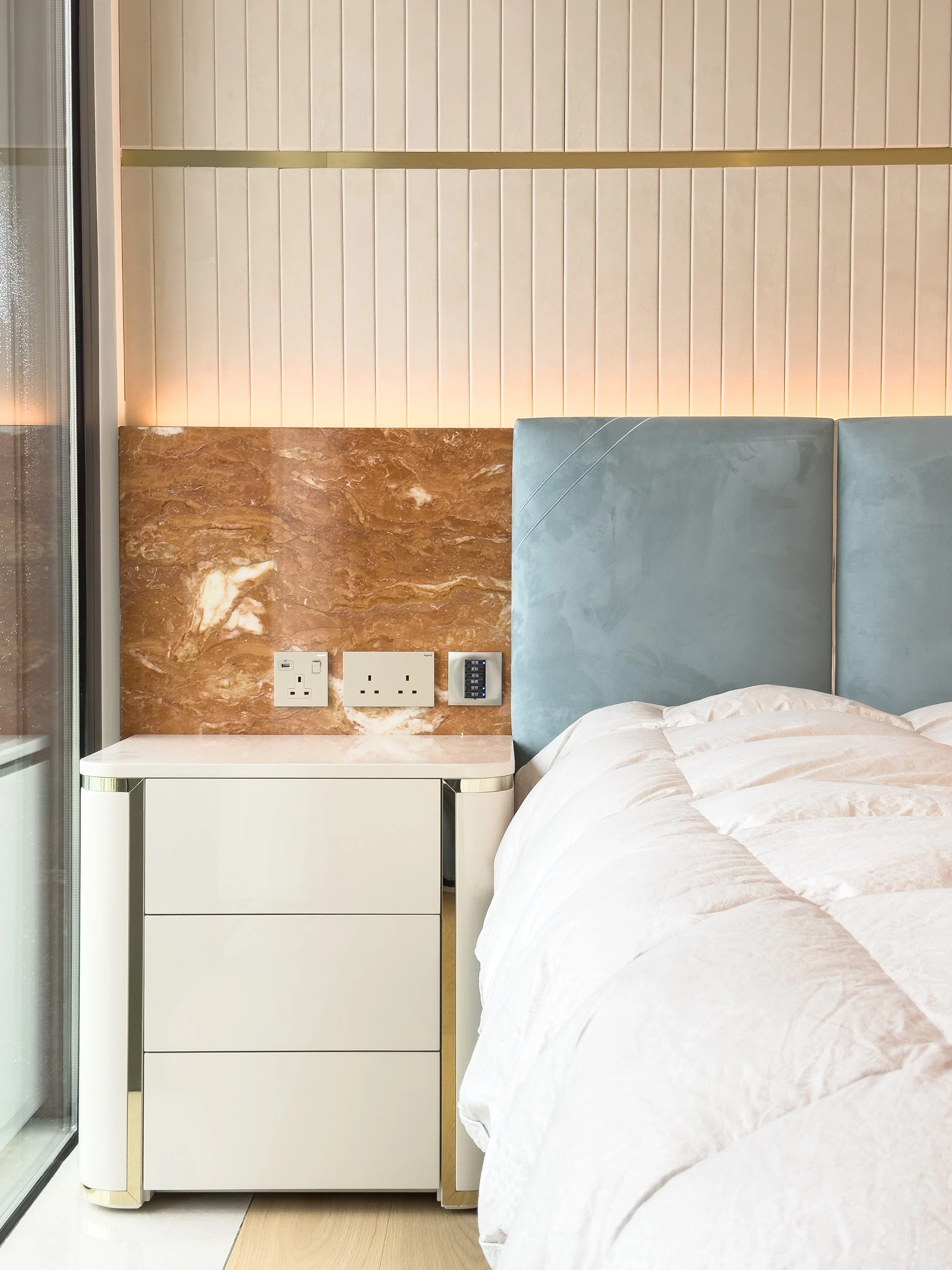 Close-up of a modern hotel bedroom nightstand with power outlets and a phone control panel, next to a bed with a soft, quilted white blanket and a padded light blue headboard, against a wall with warm lighting and wood-paneled accents.