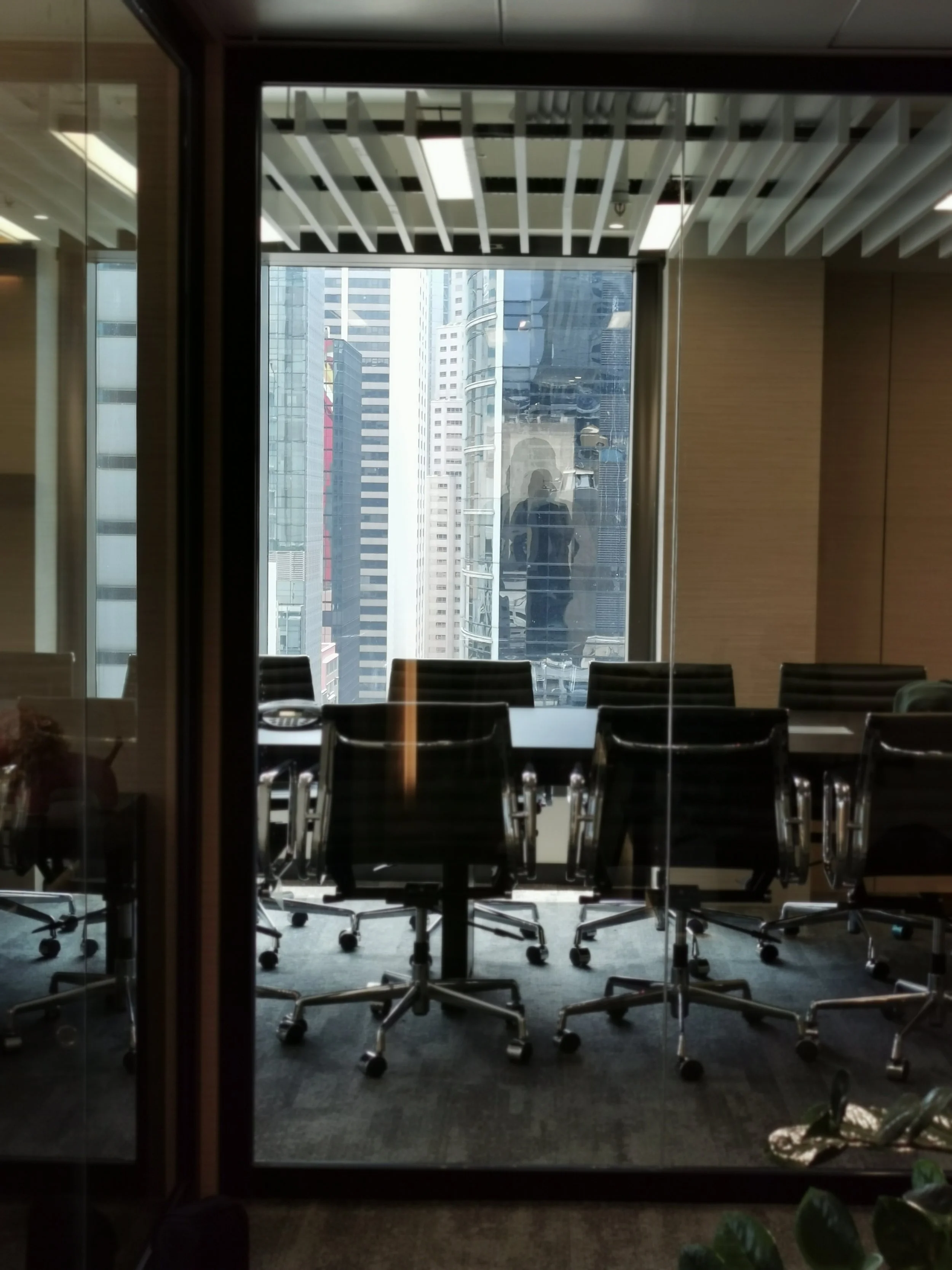 Conference room with black office chairs and a large window showing a cityscape of tall buildings, with a person reflected in the window.