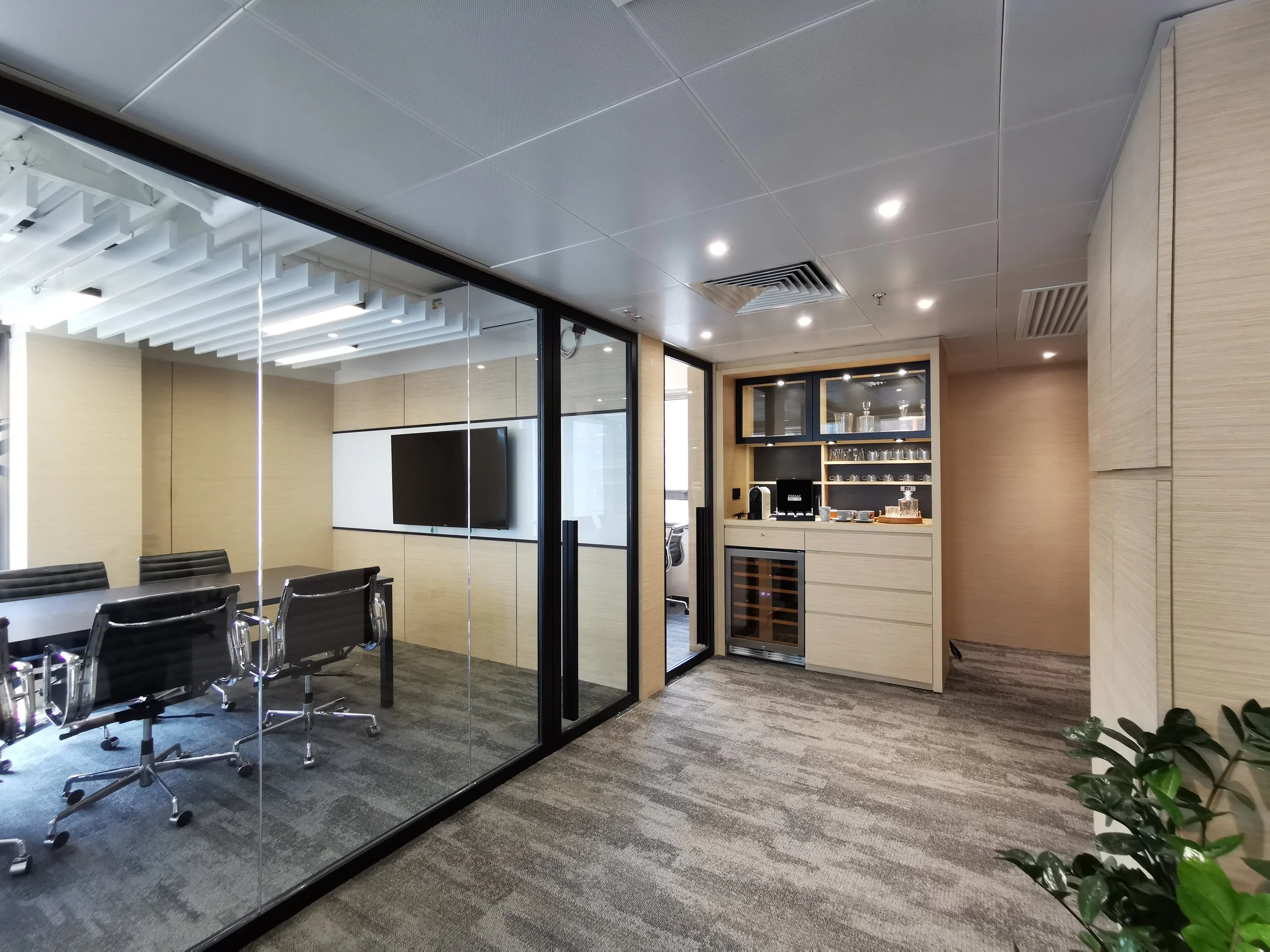 Modern office conference room with glass walls, black conference table and chairs, a wall-mounted TV, and a kitchenette area with glassware and a wine cooler.