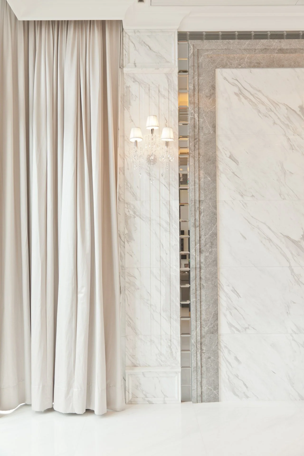 Interior wall with marble paneling, mirrored shelving, elegant wall sconce with lampshades, and beige curtains.