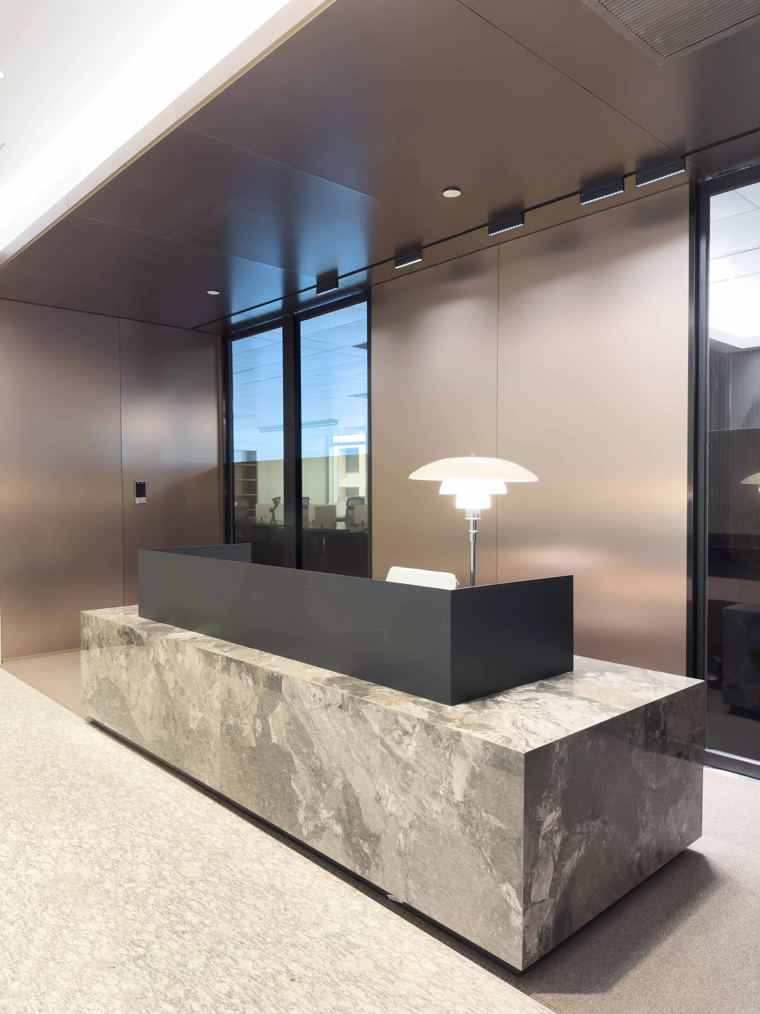 Modern reception desk made of marble with a sleek black partition, illuminated by a white desk lamp, in a contemporary office lobby with glass walls and minimalistic design.