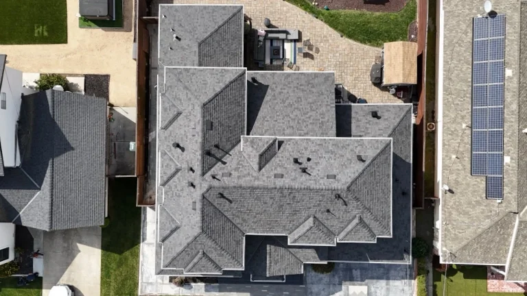 An aerial view of a residential house with a gray rooftops, neighboring houses, a solar panel installation on a neighboring roof, and a backyard.