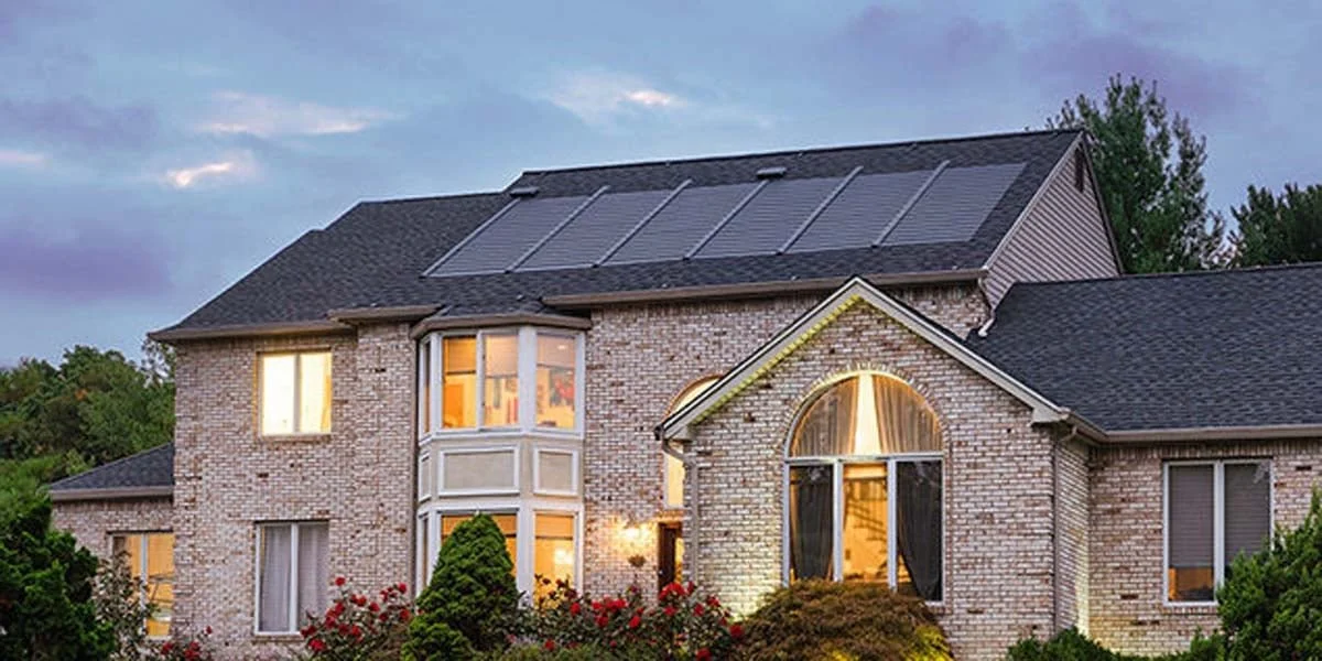 Two-story brick house with solar panels on the roof, illuminated windows, and a well-maintained front yard with bushes and flowers, during twilight.