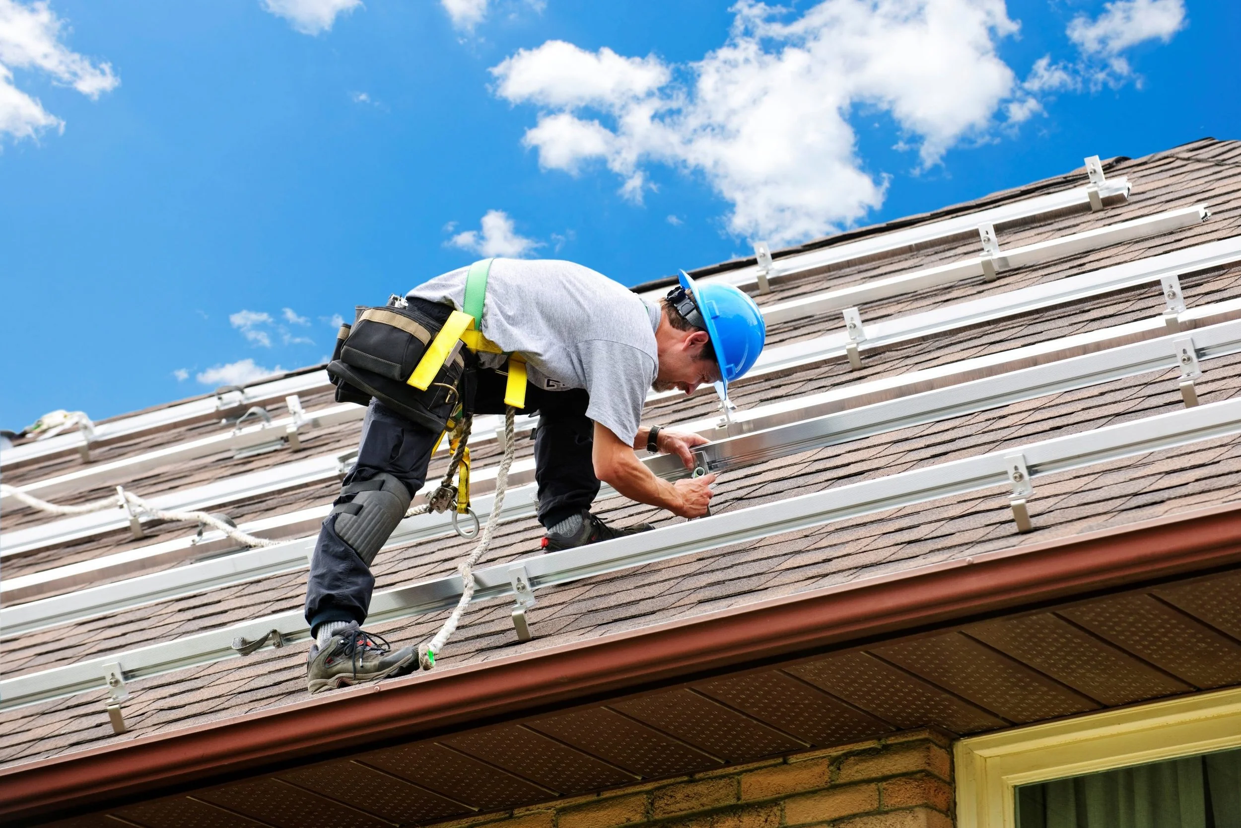 A worker wearing a blue safety helmet and harness is installing or repairing solar panels on the roof of a house on a bright, sunny day with a blue sky and some clouds.