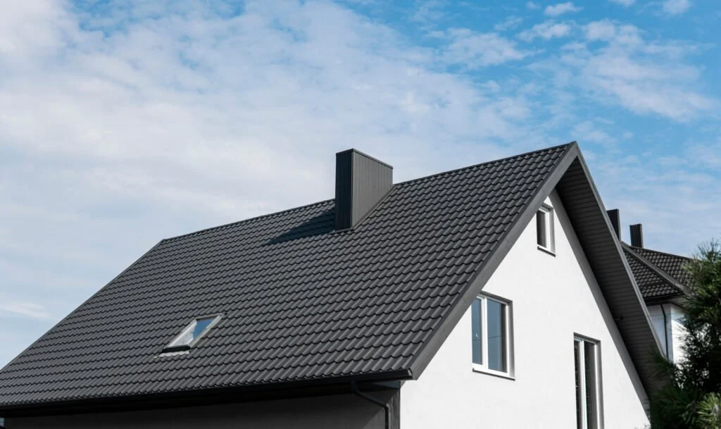 Front view of a modern house with a grey tiled steep roof, white walls, and several windows under a partly cloudy sky.