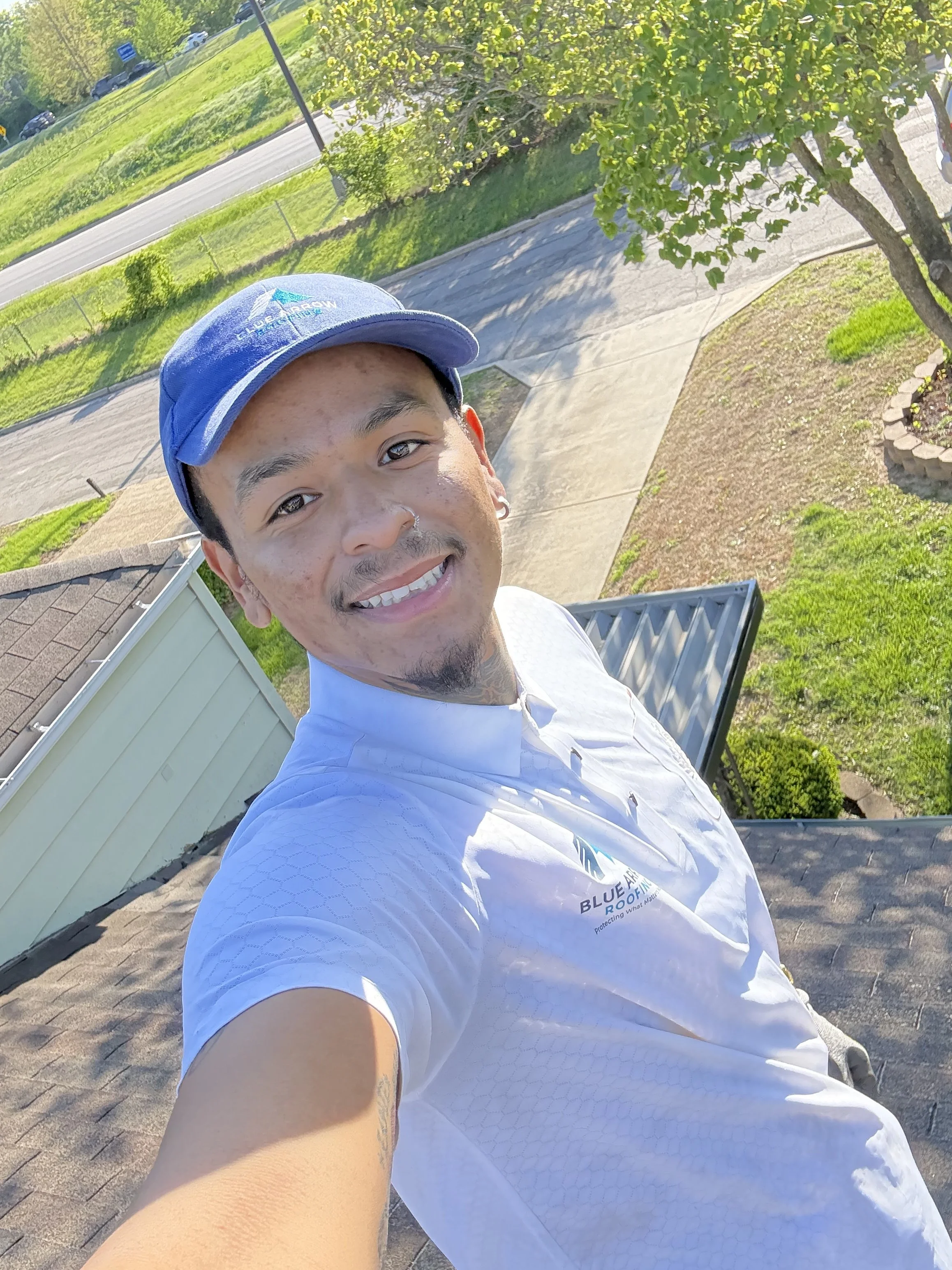 A young man taking a selfie outdoors in daytime, wearing a blue cap and a white athletic shirt, with grass, trees, pathway, and parked cars in the background.