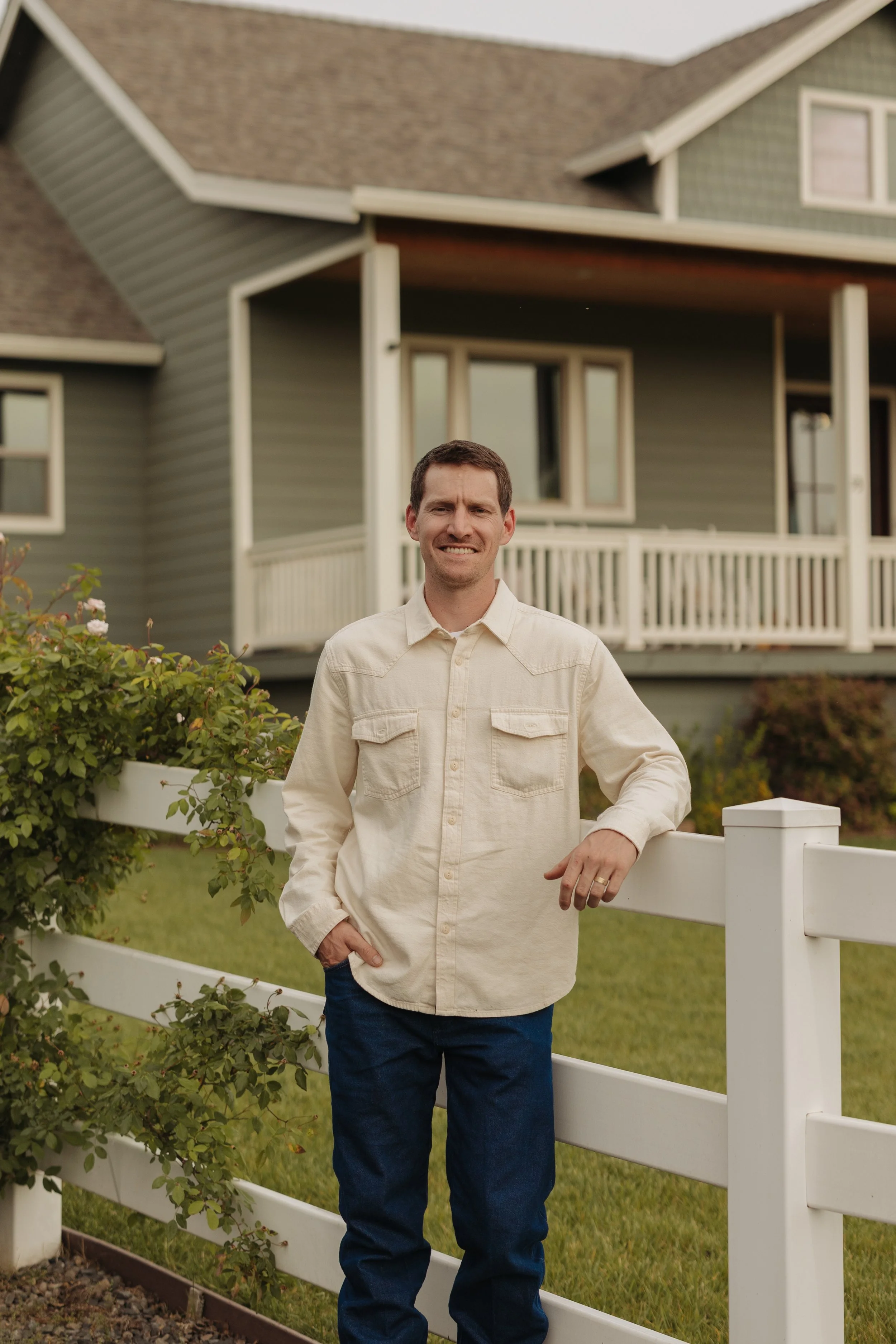 A man standing by a white fence in front of a house with a green exterior and a large porch, smiling and wearing a light-colored shirt and dark pants.