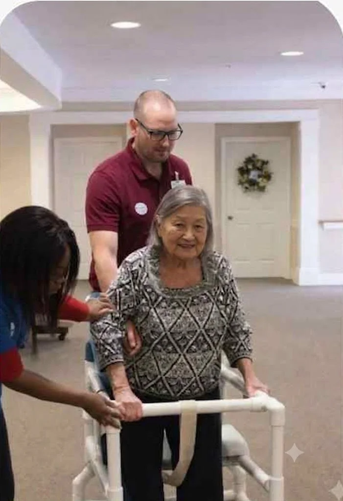 An elderly Asian woman smiling while using a walker, supported by two caring healthcare staff members in an assisted living facility.