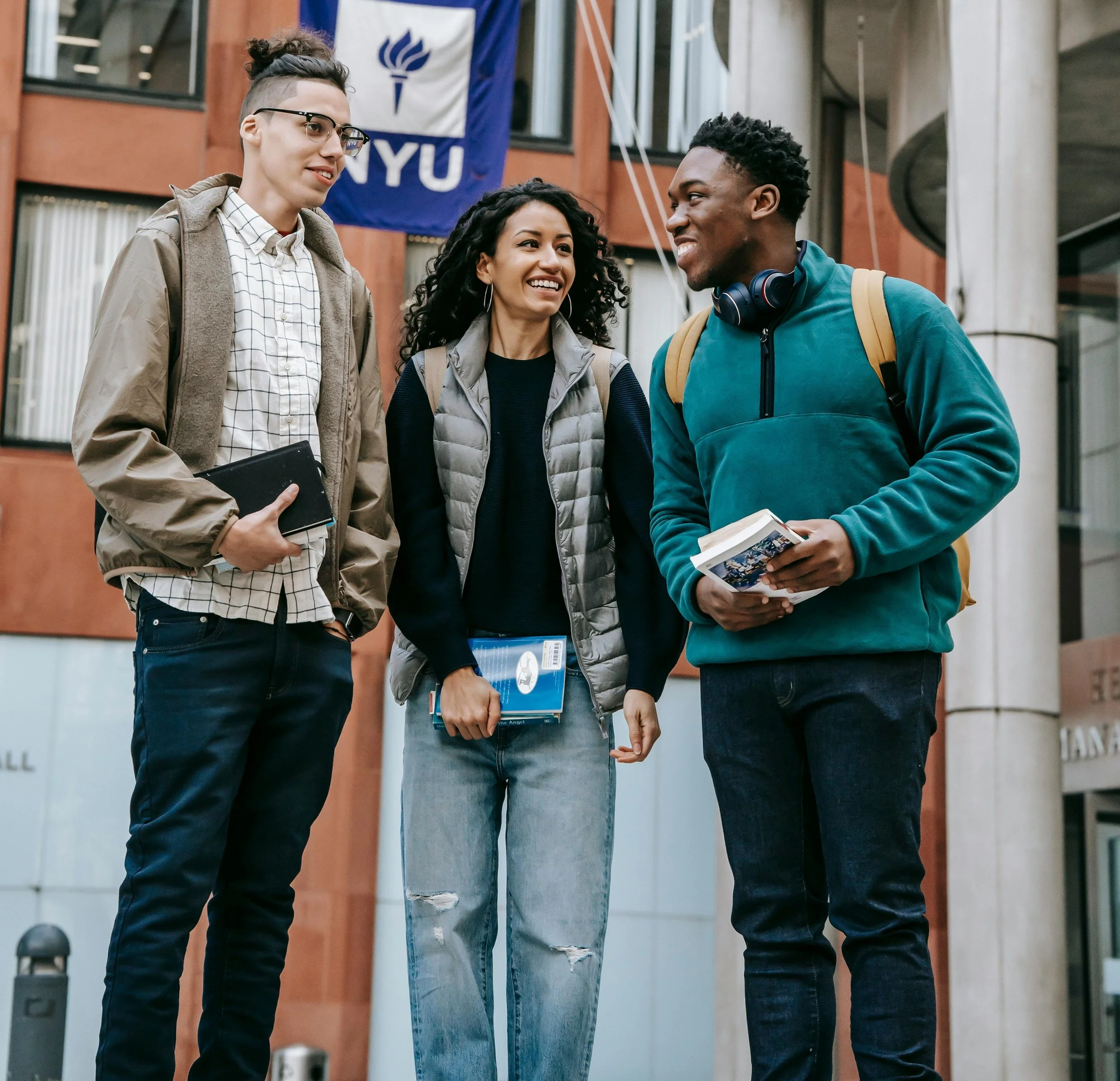 Three college students standing outside NYU building, talking and smiling, holding books and notebooks.