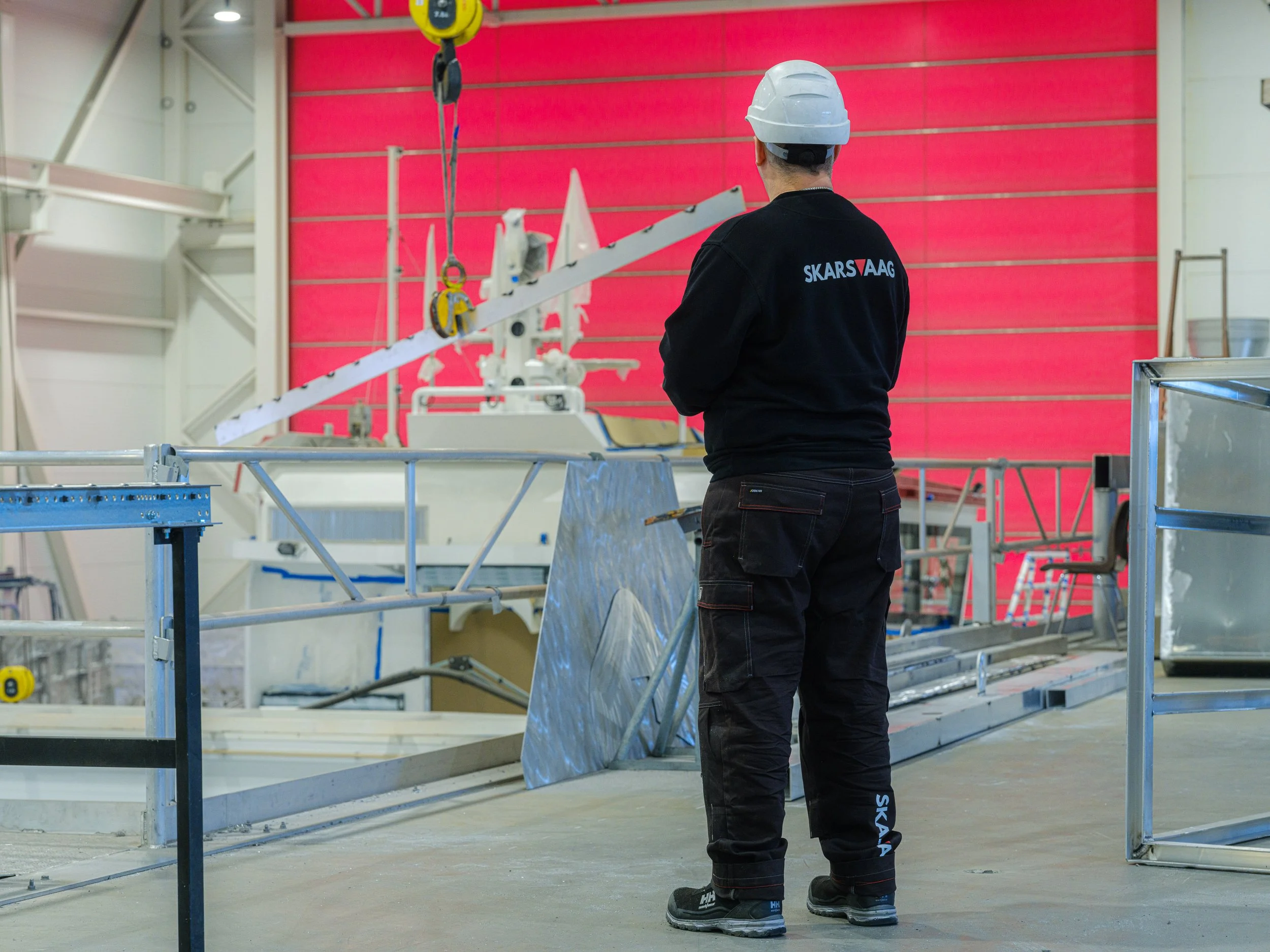 En person iført svart arbeidsantrekk med teksten "SKARSVAAG" på ryggen, og verneutstyr, står i en fabrikk eller verksted hvor det pågår montering eller konstruksjon av stor maskin eller utstyr.