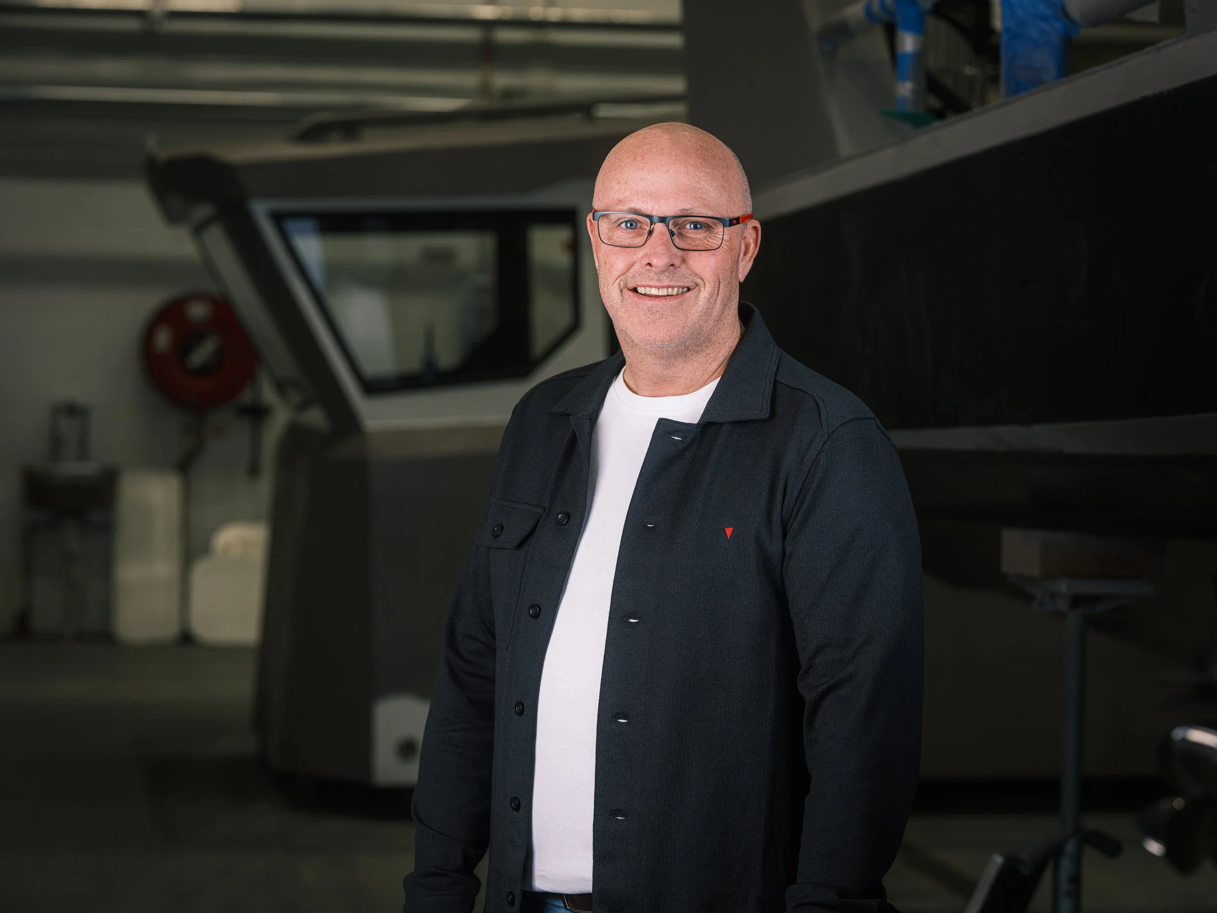 En mann med briller smiler foran en stor maskin i en industriell setting.