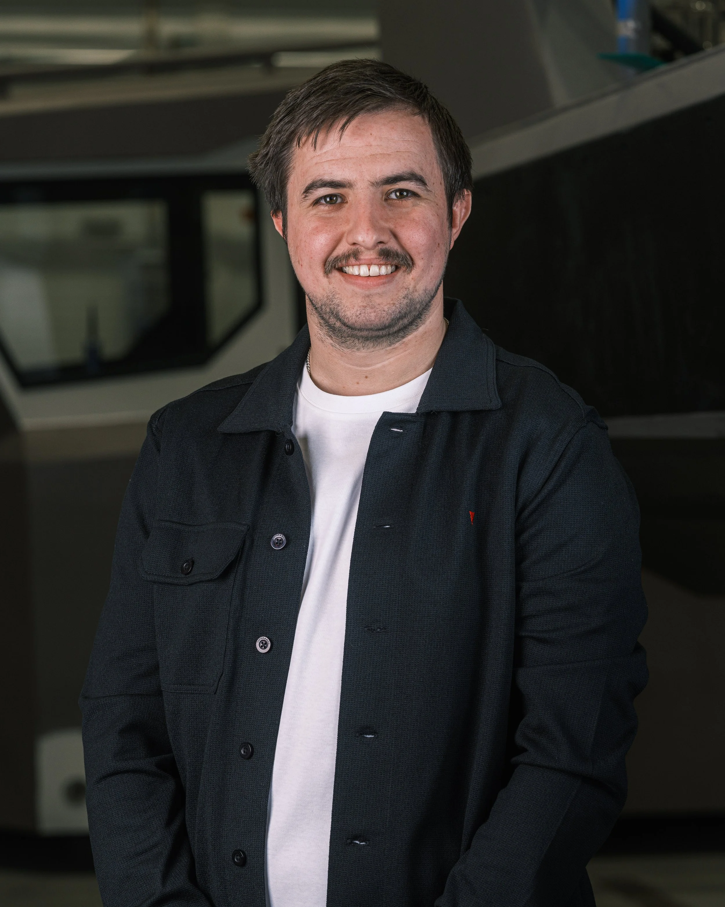 Portrett av en mann med kort brunt hår, bustet skjegg og stakkars smil, iført en svart jakke og hvit T-skjorte. Bakgrunn med noe utstyr og svart vegg.