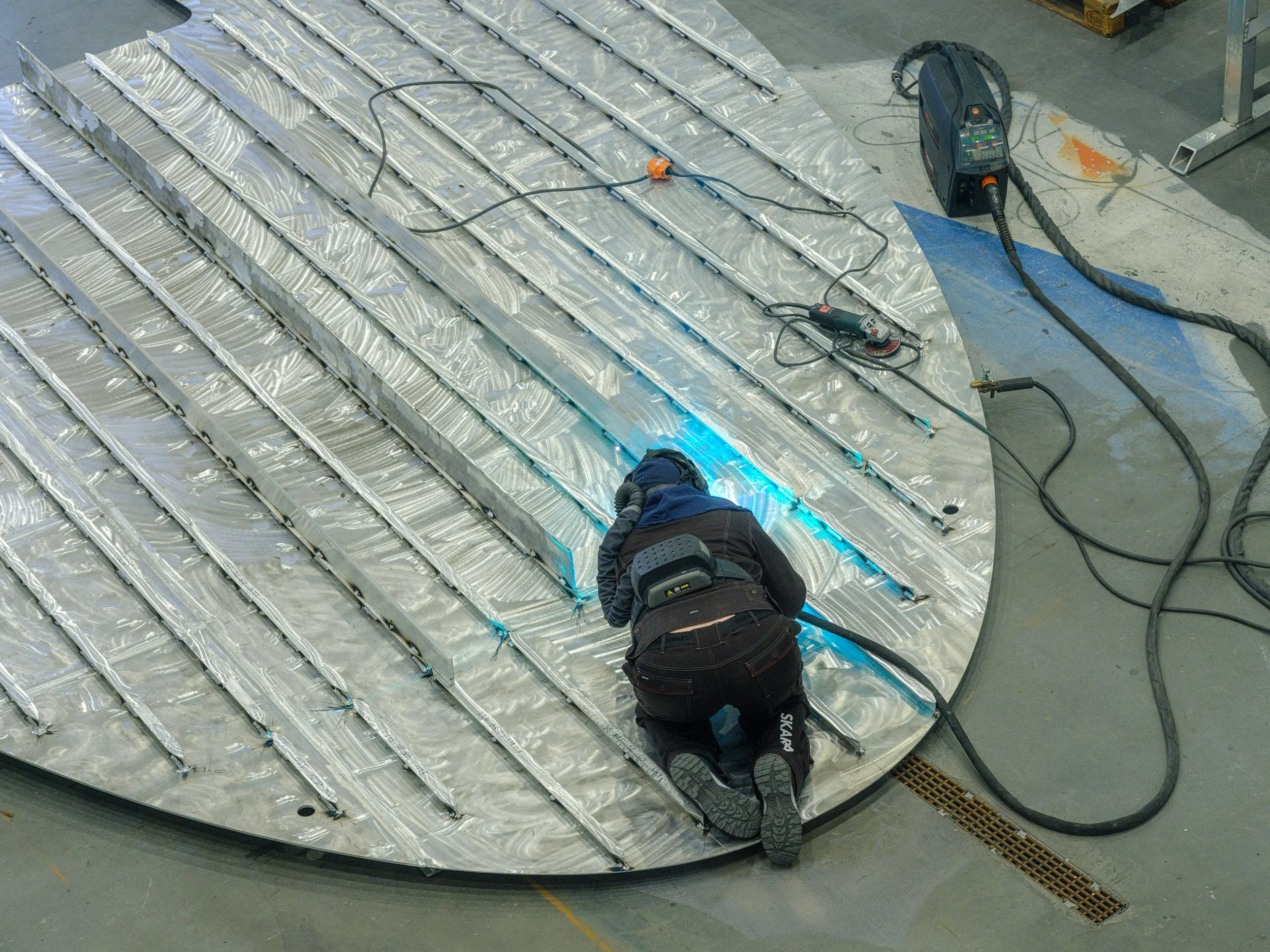 To personer jobber med å installere eller reparere et metallpanel på gulvet i en industribygg, omgitt av verktøy og utstyr.