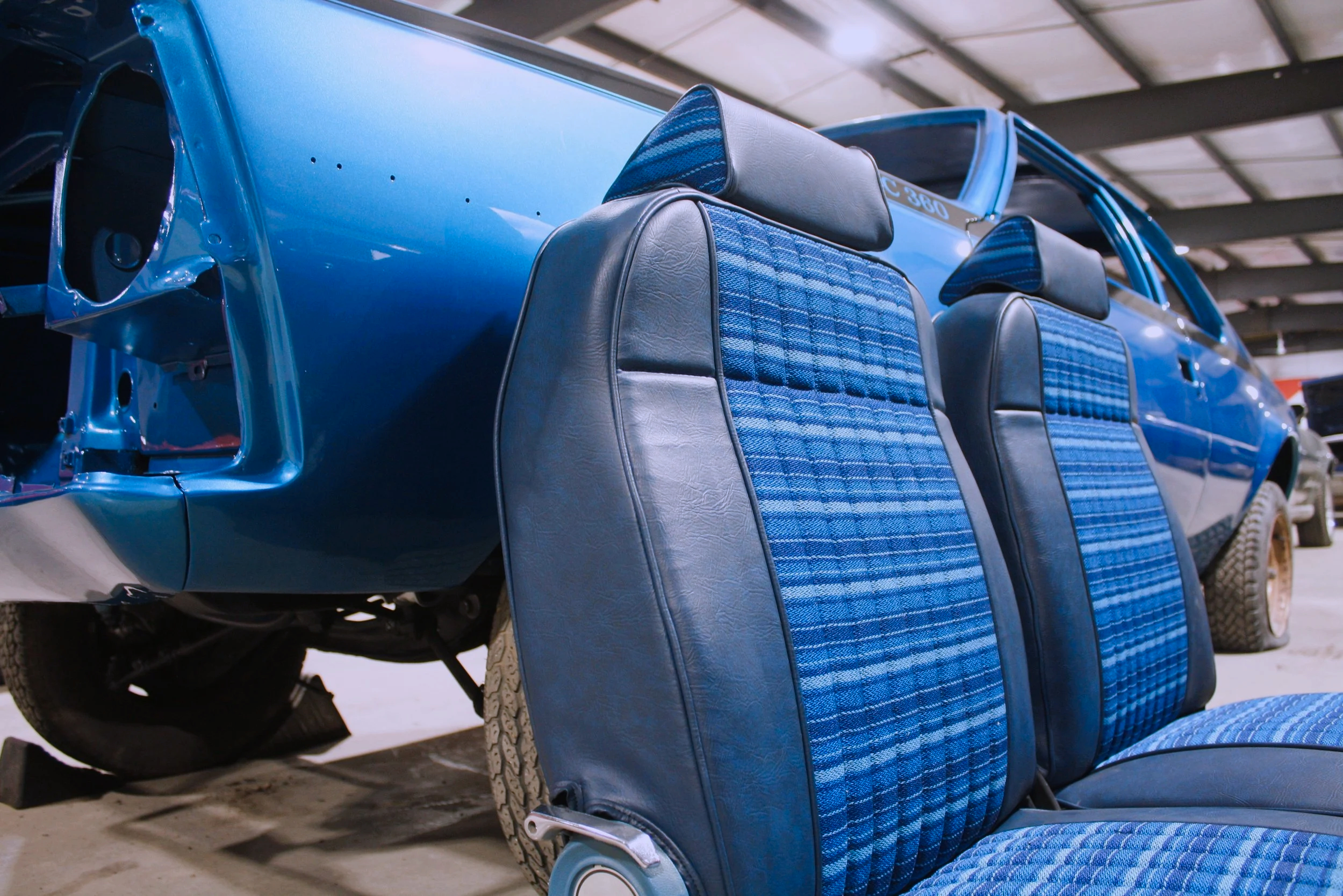 Blue vintage car with its front panel missing, showing the inner structure, parked indoors. Two blue and black fabric and leather car seats are in the foreground.
