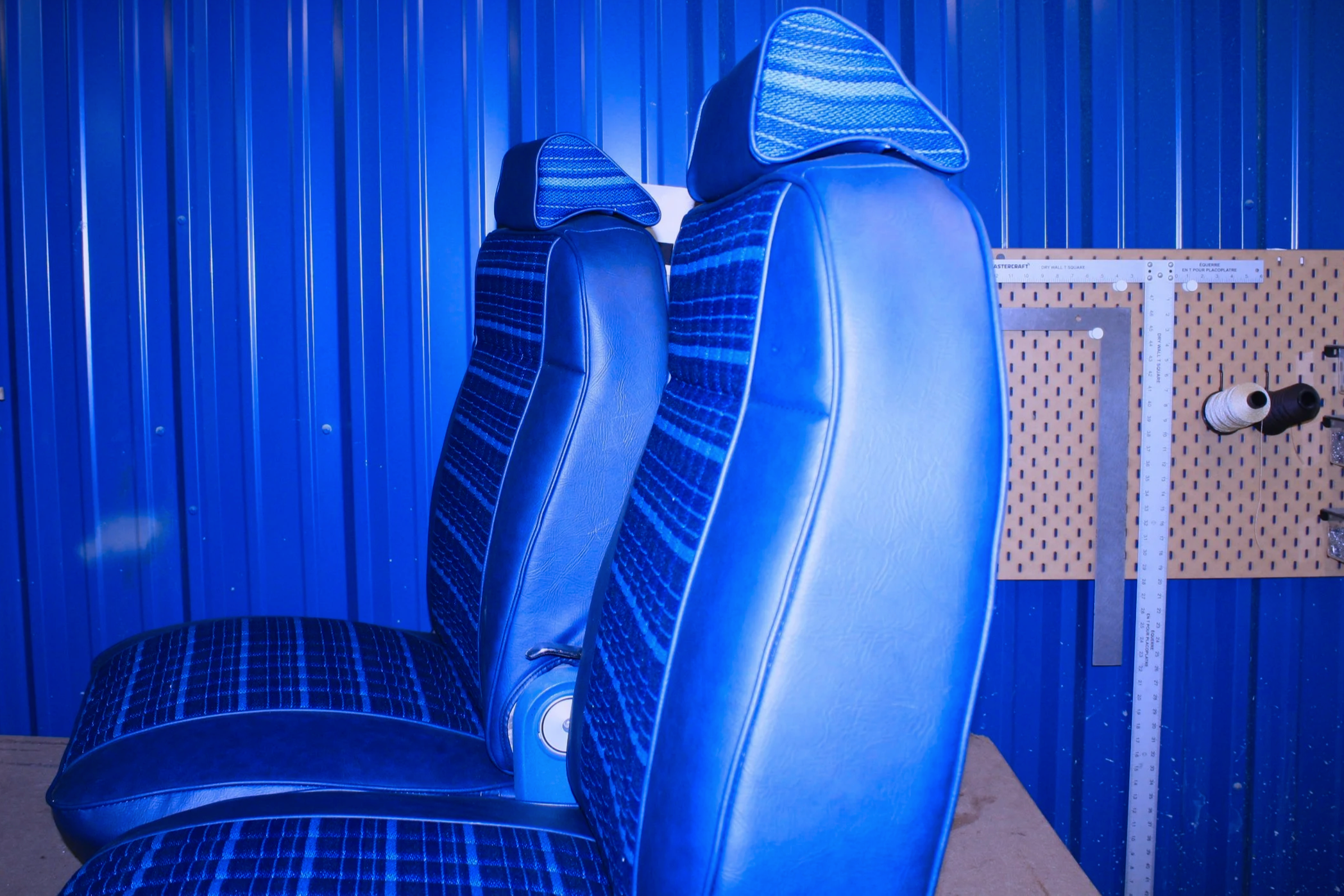 Two blue bus seats with headrests and fabric upholstery, set against a blue metal wall, with a pegboard in the background holding spools of thread.