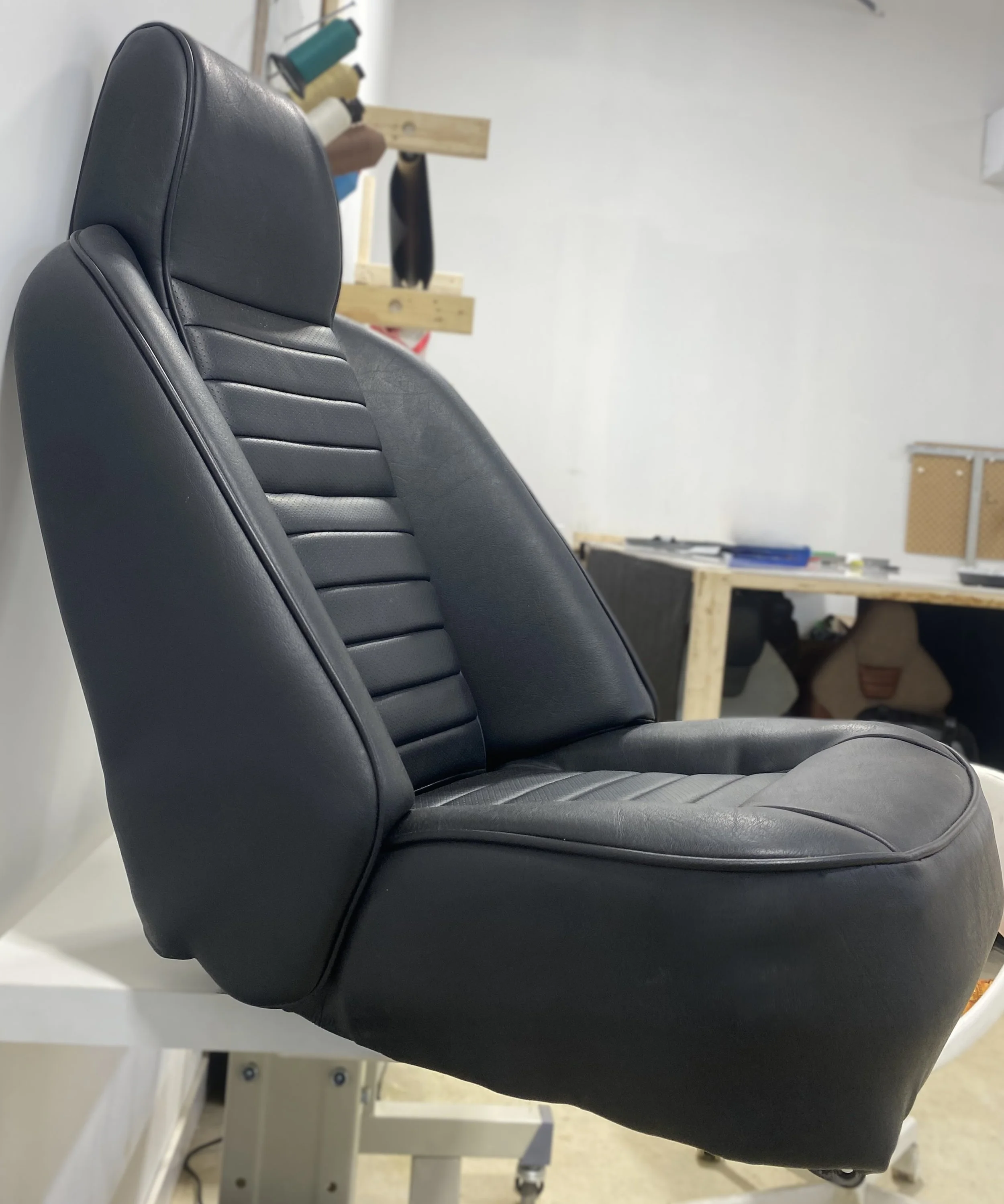 A black leather car seat with horizontal stitching, placed on a white worktable in a workshop setting.