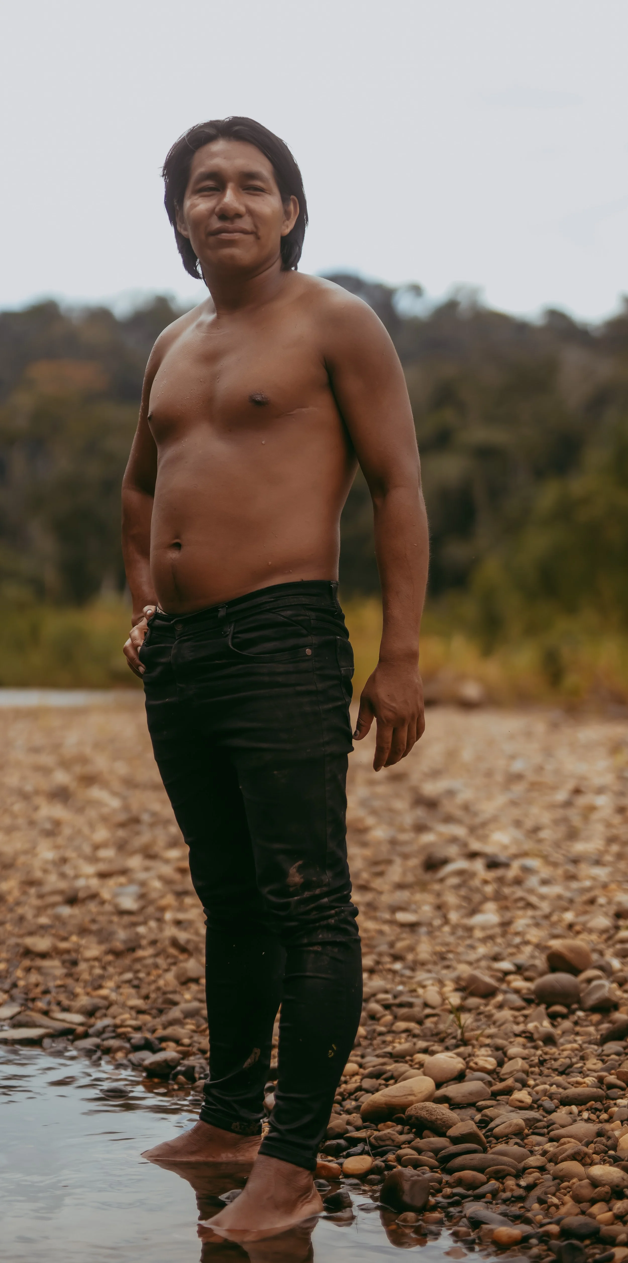 A shirtless man standing in shallow water on a rocky riverbank with a blurred natural landscape background.