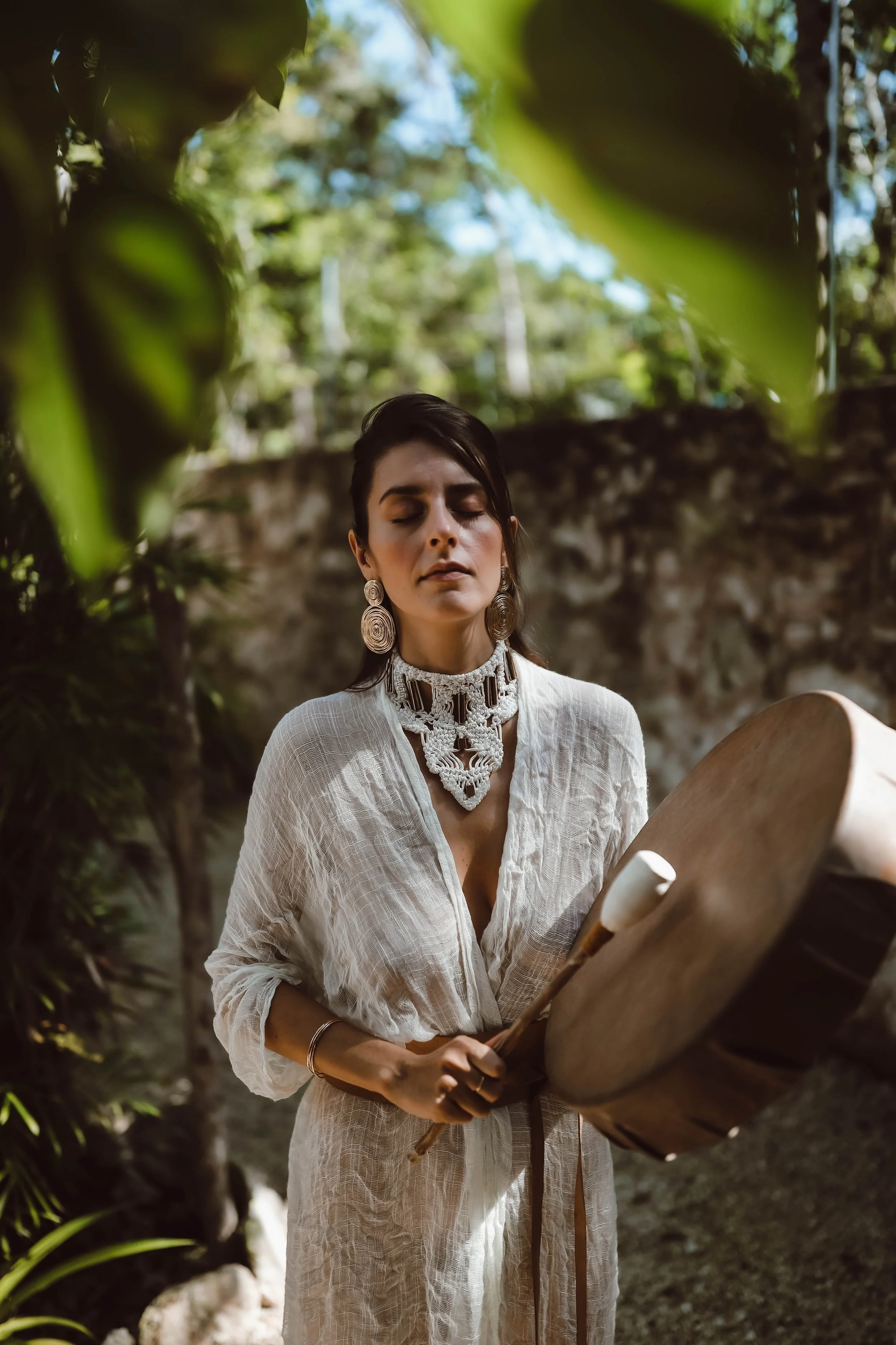 A woman in a white, textured dress with large earrings and an intricate necklace plays a large drum outdoors surrounded by green foliage.