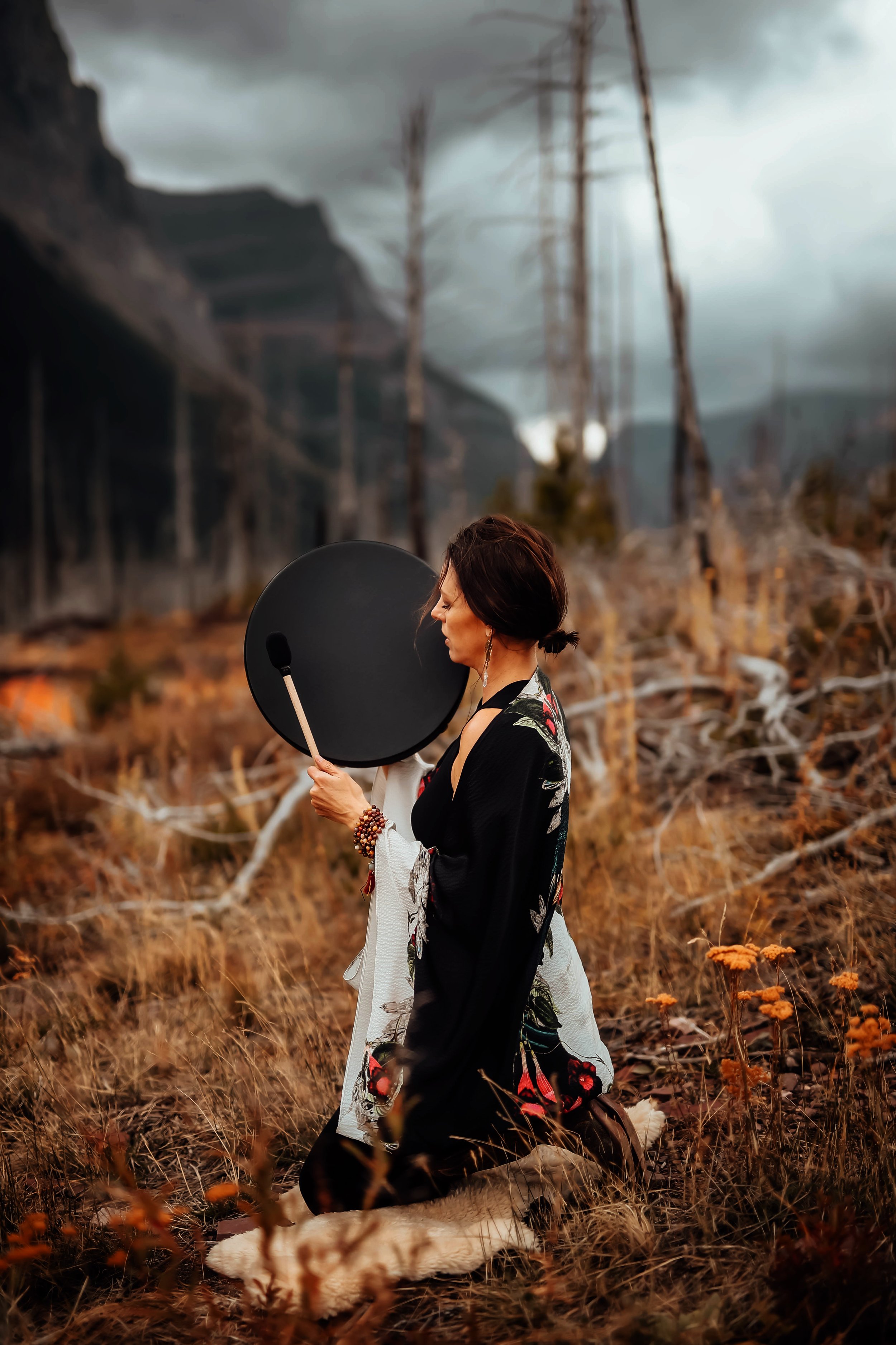 A woman kneeling in a forest clearing during dusk, holding a black small handheld gong near her face, wearing a decorated cloak with floral patterns and jewelry, surrounded by dried grass and wildflowers with a dark, cloudy sky overhead.