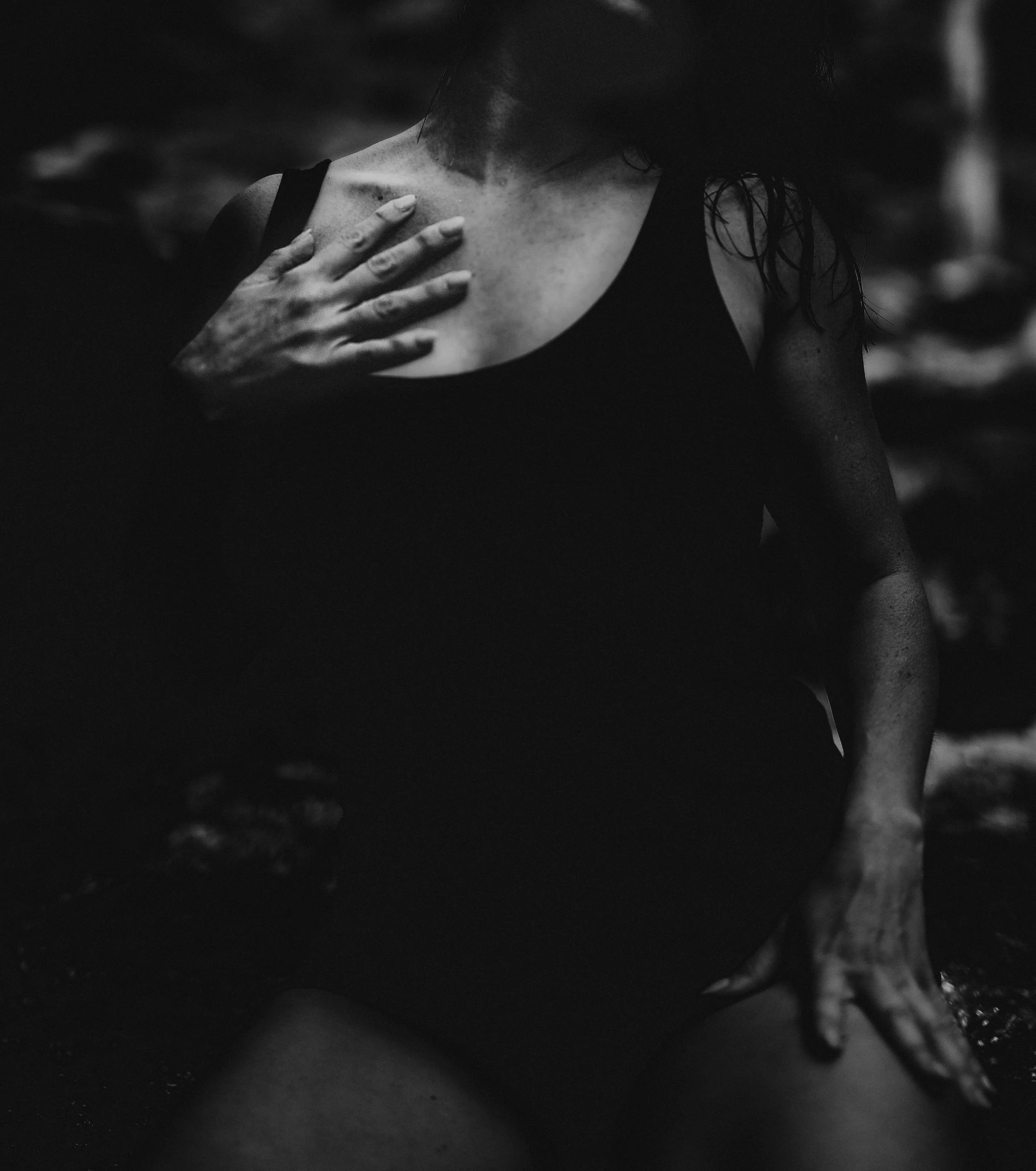 Black and white photo of a woman with wet hair wearing a sleeveless top, touching her chest with her left hand, in a dark setting.
