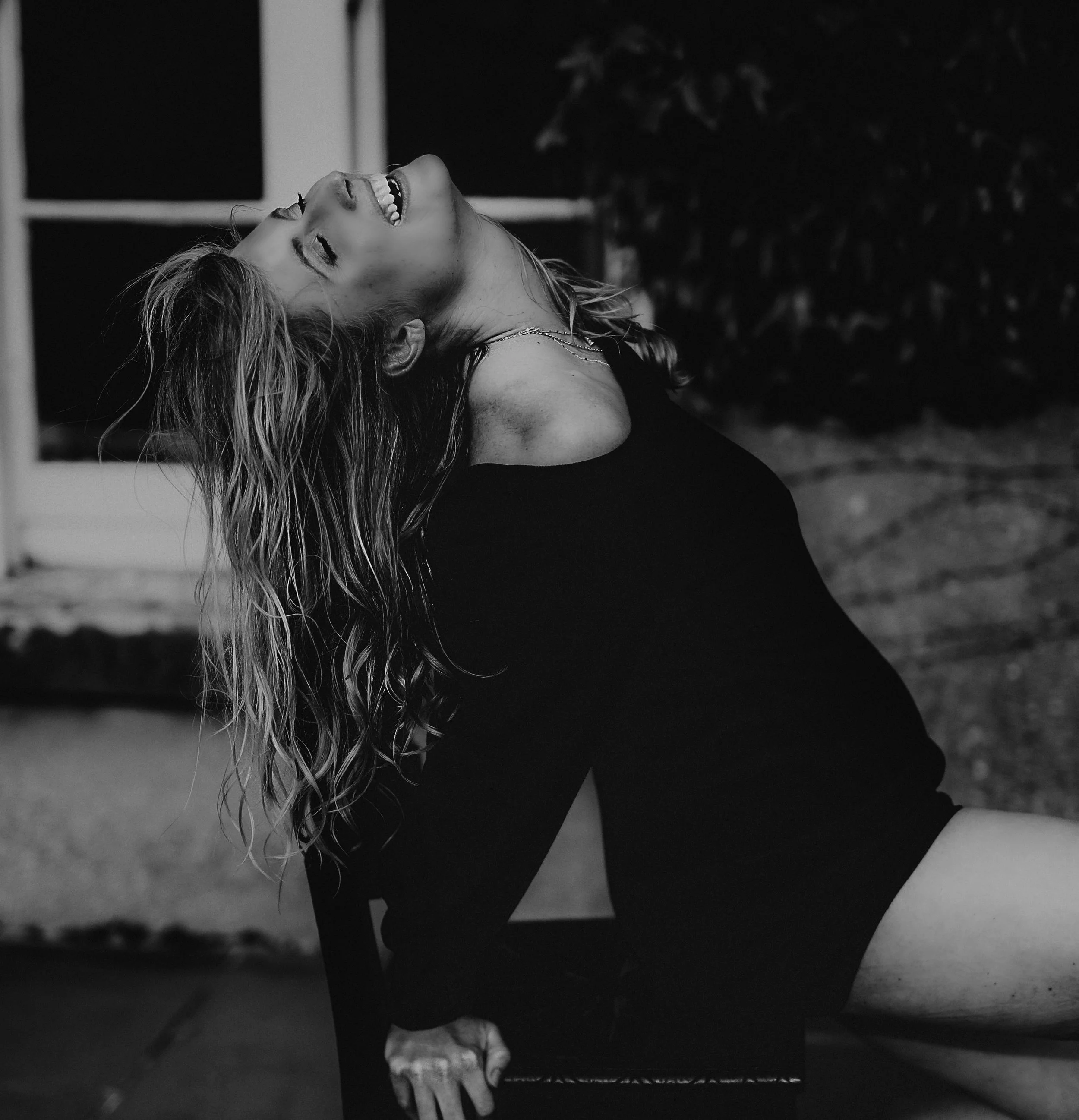 A woman with wavy hair leaning back with her head tilted and laughing in front of a window at night.