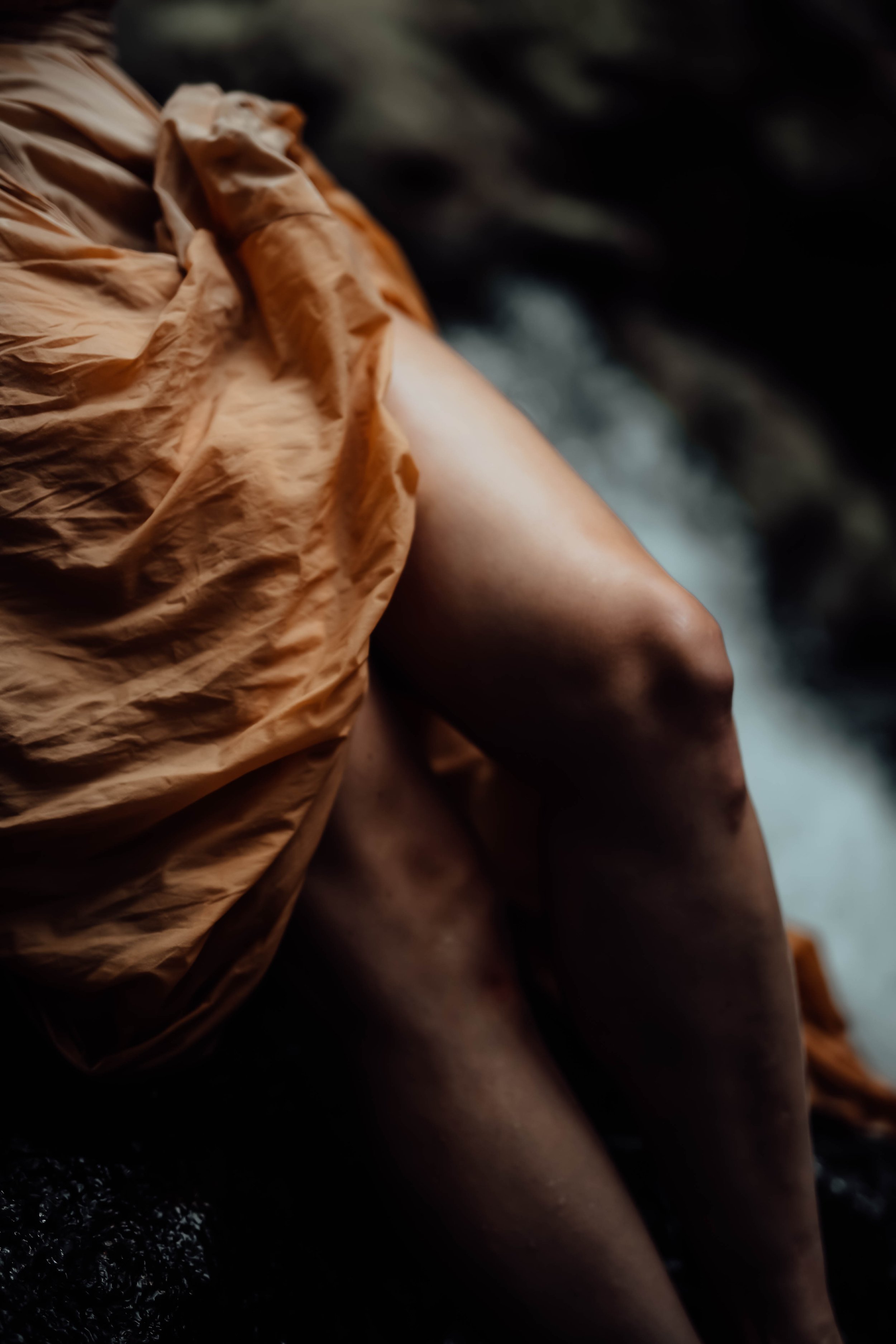 Close-up of a person's leg and thigh peeking out from a brown, wrinkled fabric with a blurred natural background.