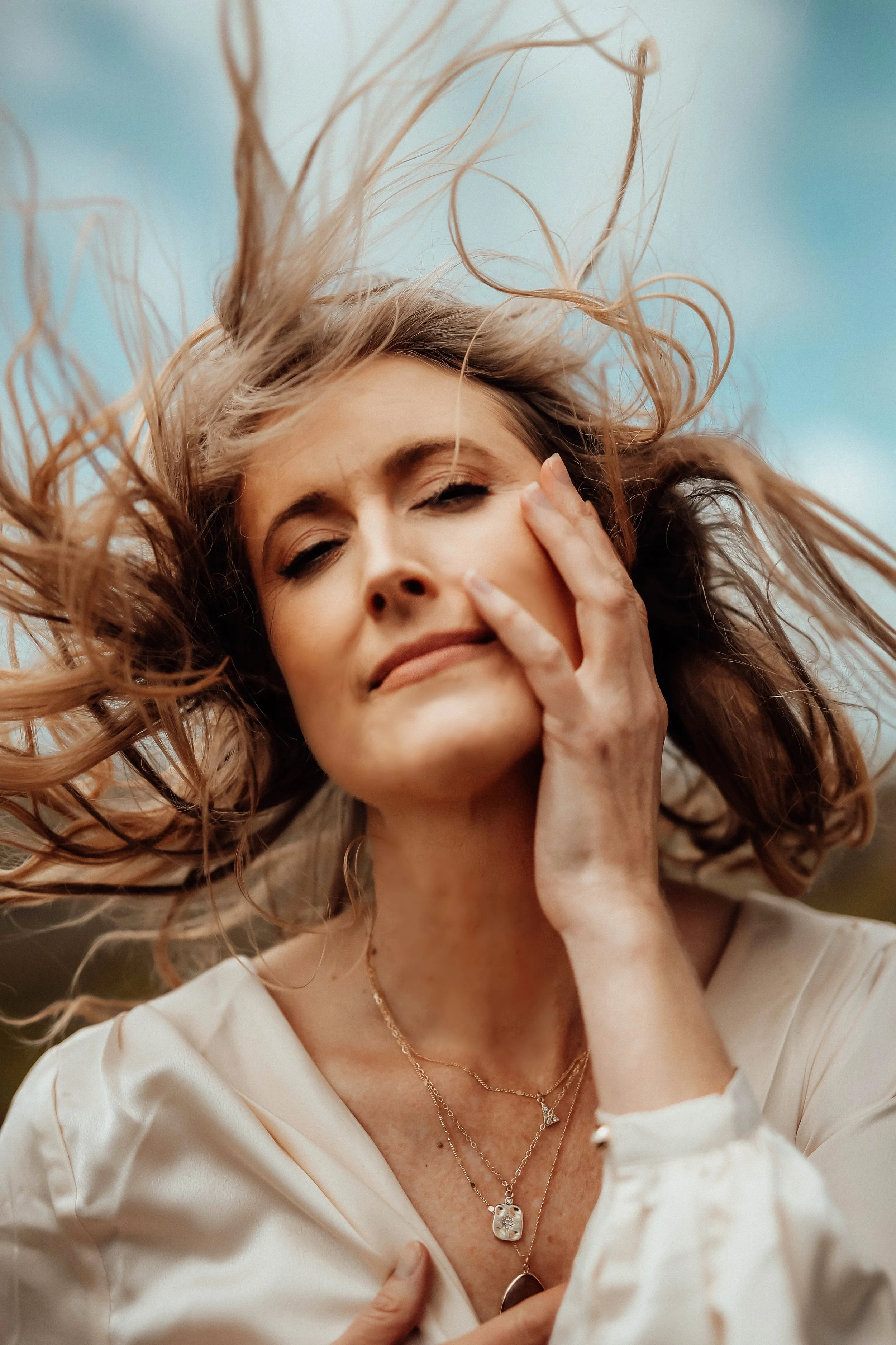 A woman with wavy hair and multiple necklaces, one with a star pendant, tilting her head slightly with a serene expression against a cloudy sky.