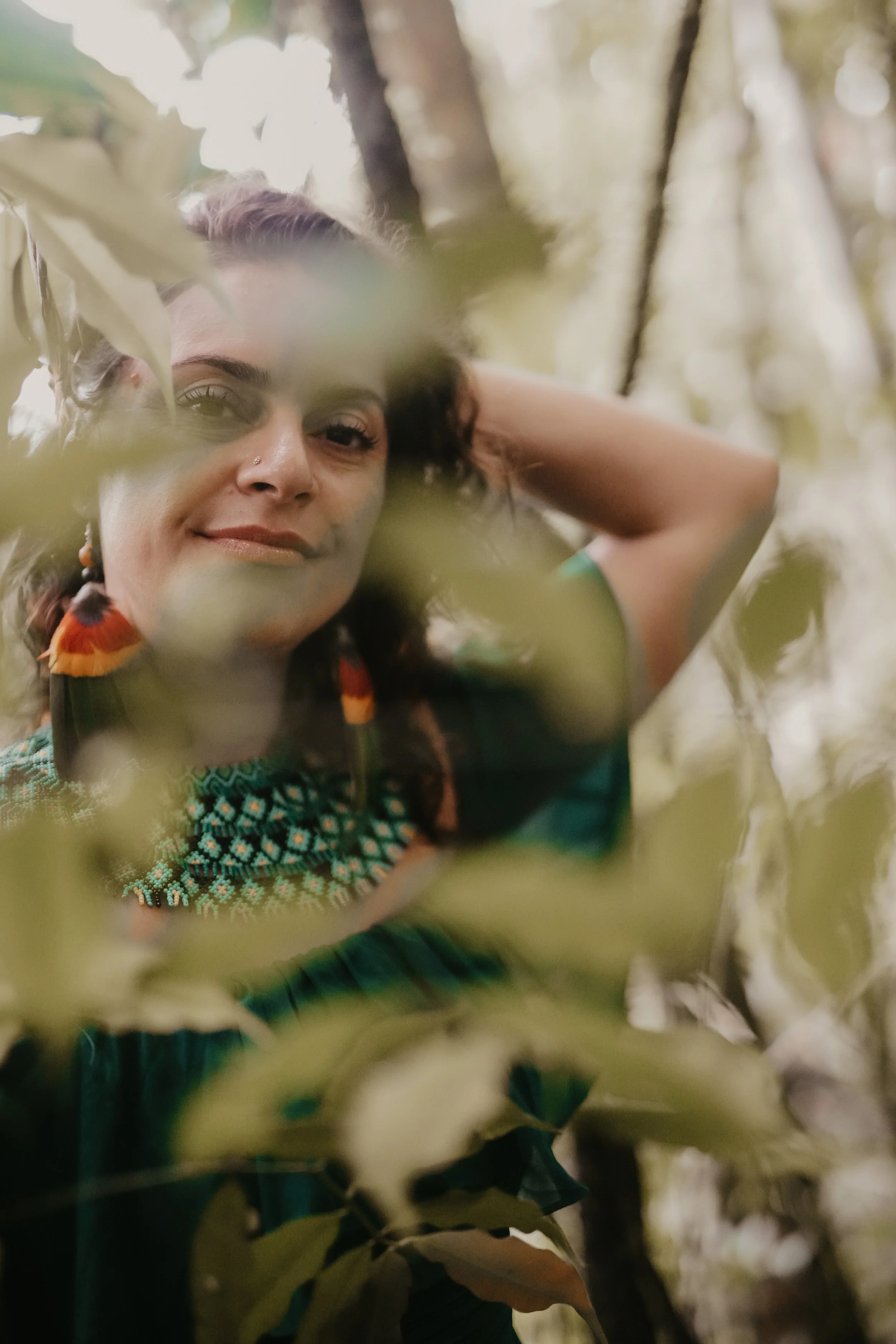 A woman with dark hair and earrings stands amid trees and leaves, looking at the camera with a relaxed expression.