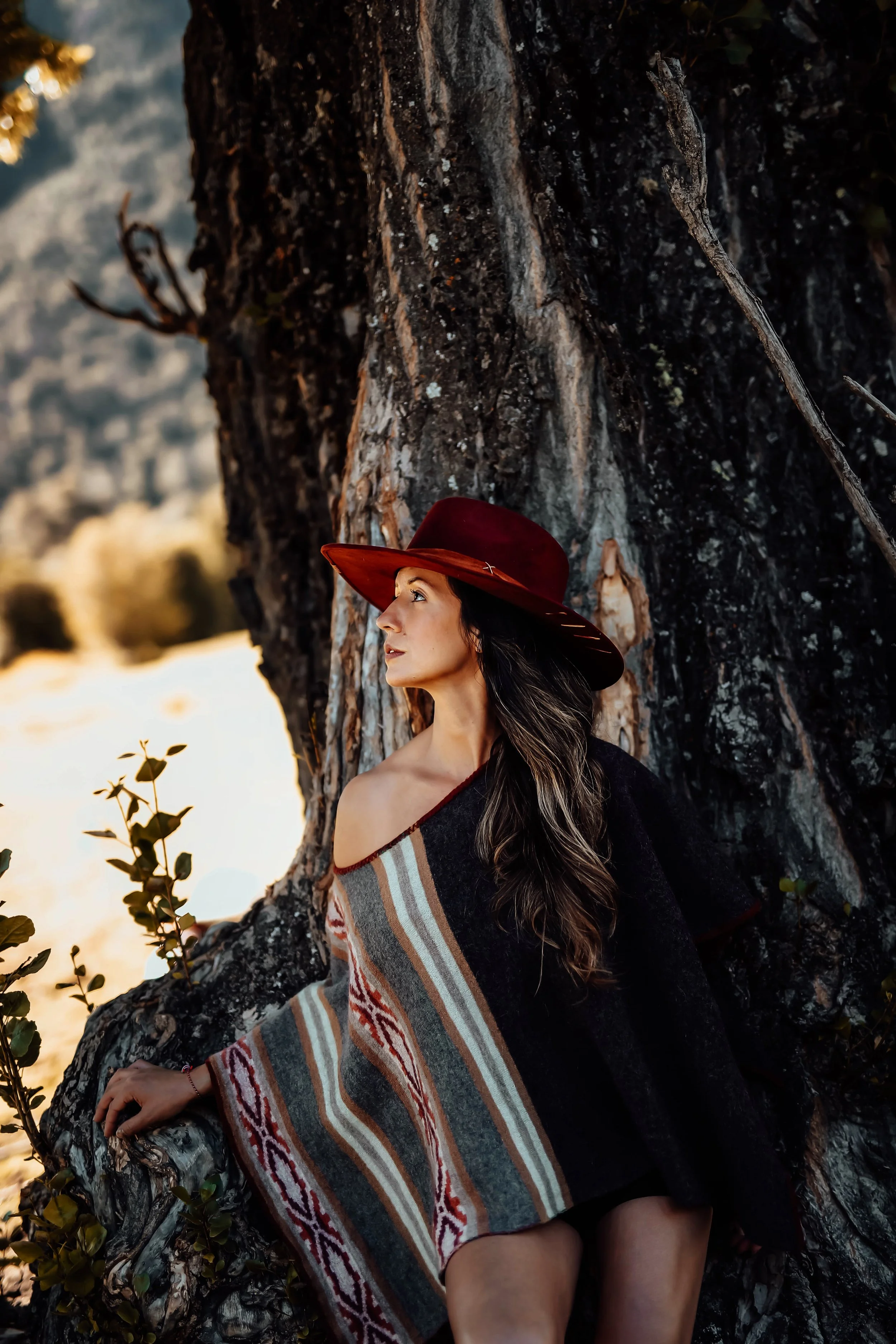 A woman with long dark hair wearing a large red hat and a patterned shawl, sitting against a large tree trunk in a natural outdoor setting.