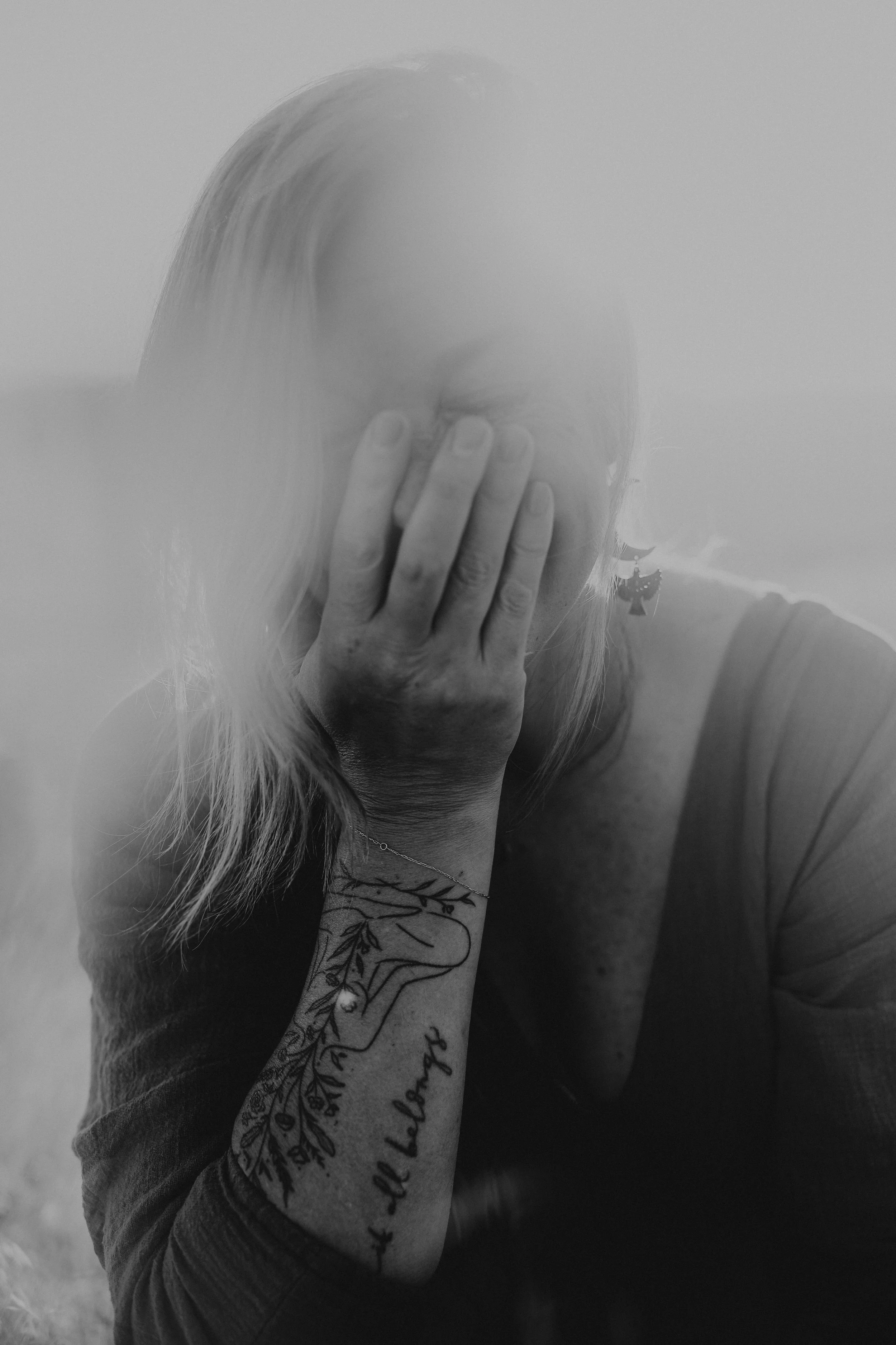 A woman with long hair covering her face, hand over her face, tattoo on her arm, wearing a dark top.