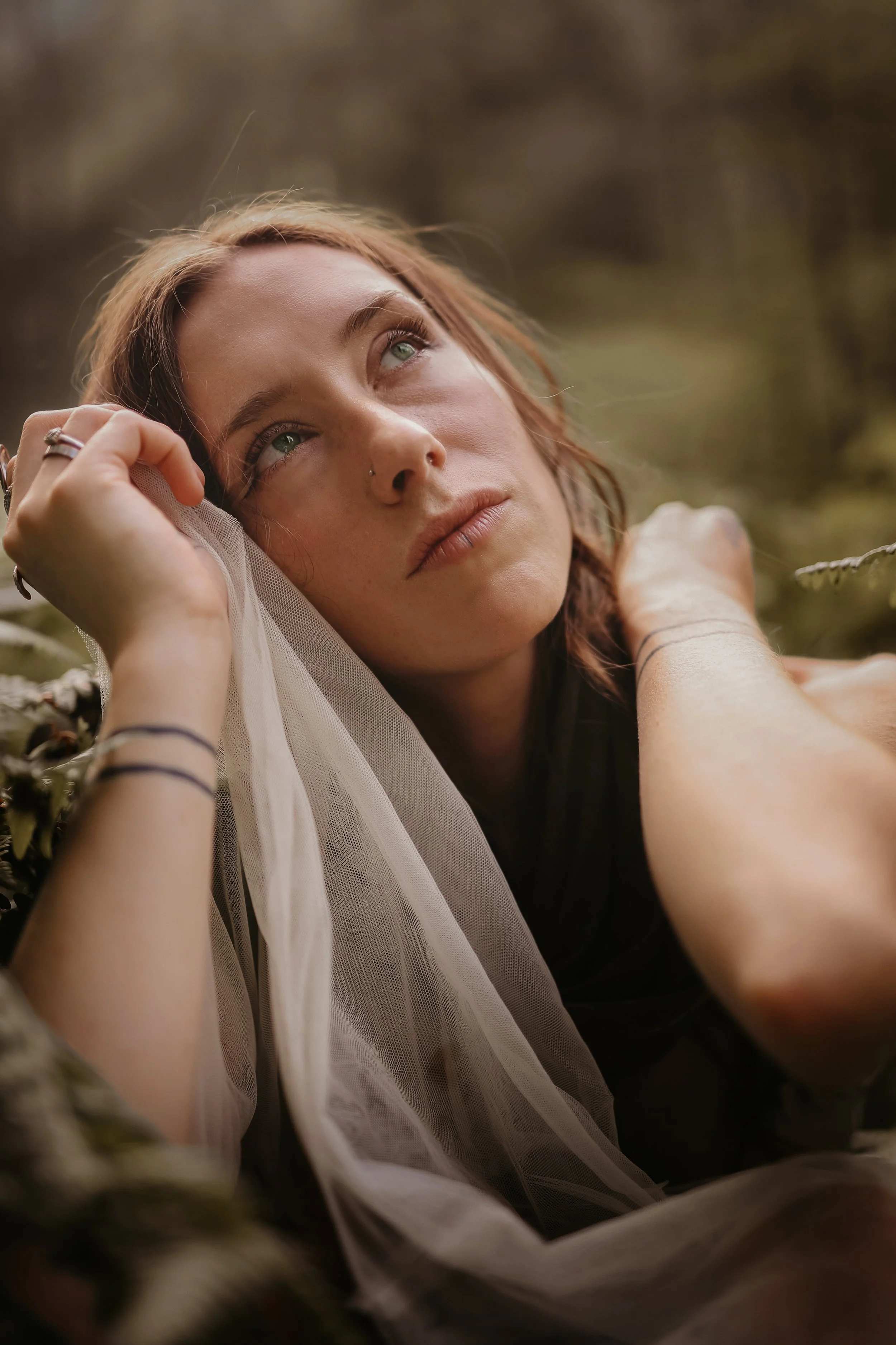 A young woman lying outdoors on a soft surface, resting her head on her hand with a contemplative expression, surrounded by natural scenery with muted earthy tones.
