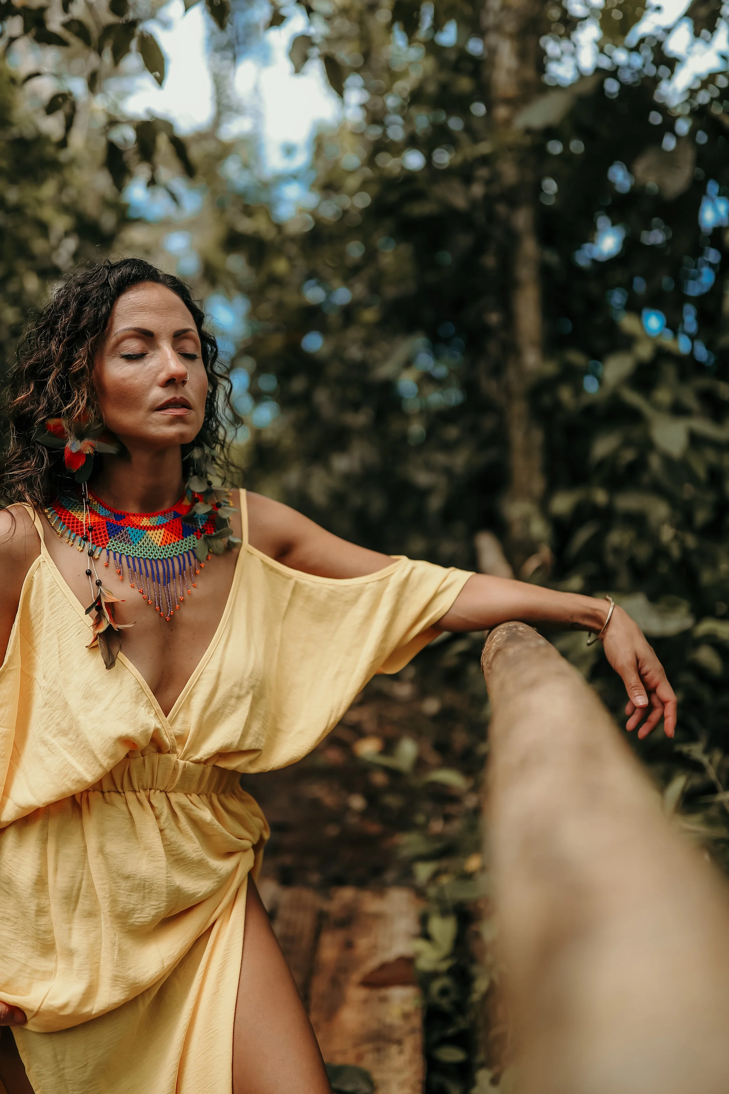 A woman with curly dark hair and tan skin, wearing a yellow off-shoulder dress and colorful beaded necklace, stands in a forest with her eyes closed, leaning on a tree branch.