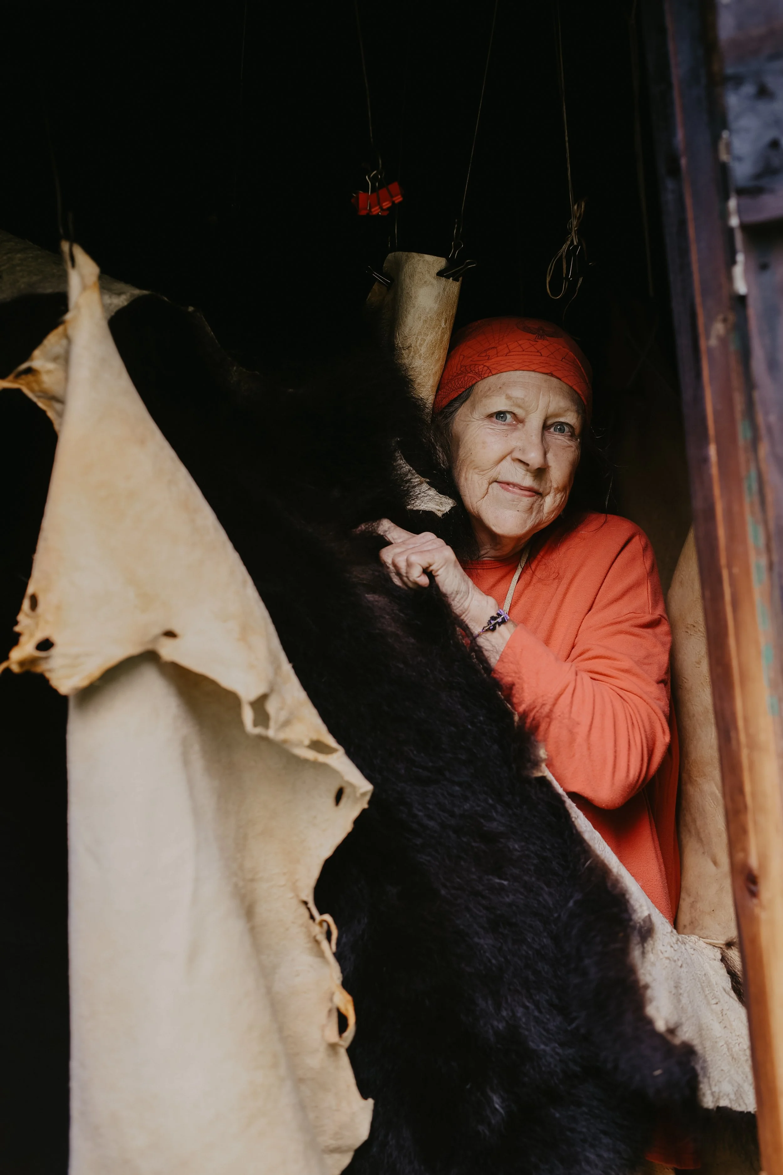 A woman in an orange headscarf and matching orange shirt standing inside a hunting cabin, holding a large black bear hide.