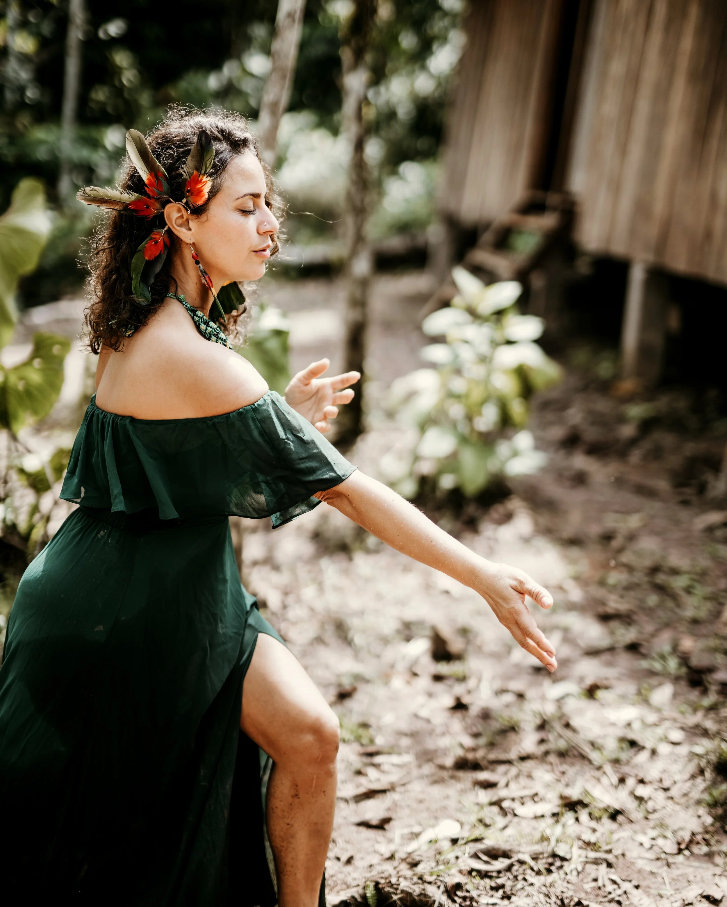 Woman dressed in a dark green off-shoulder dress with a feathered headpiece, standing outdoors in a jungle-like setting, with her eyes closed and one arm extended forward.